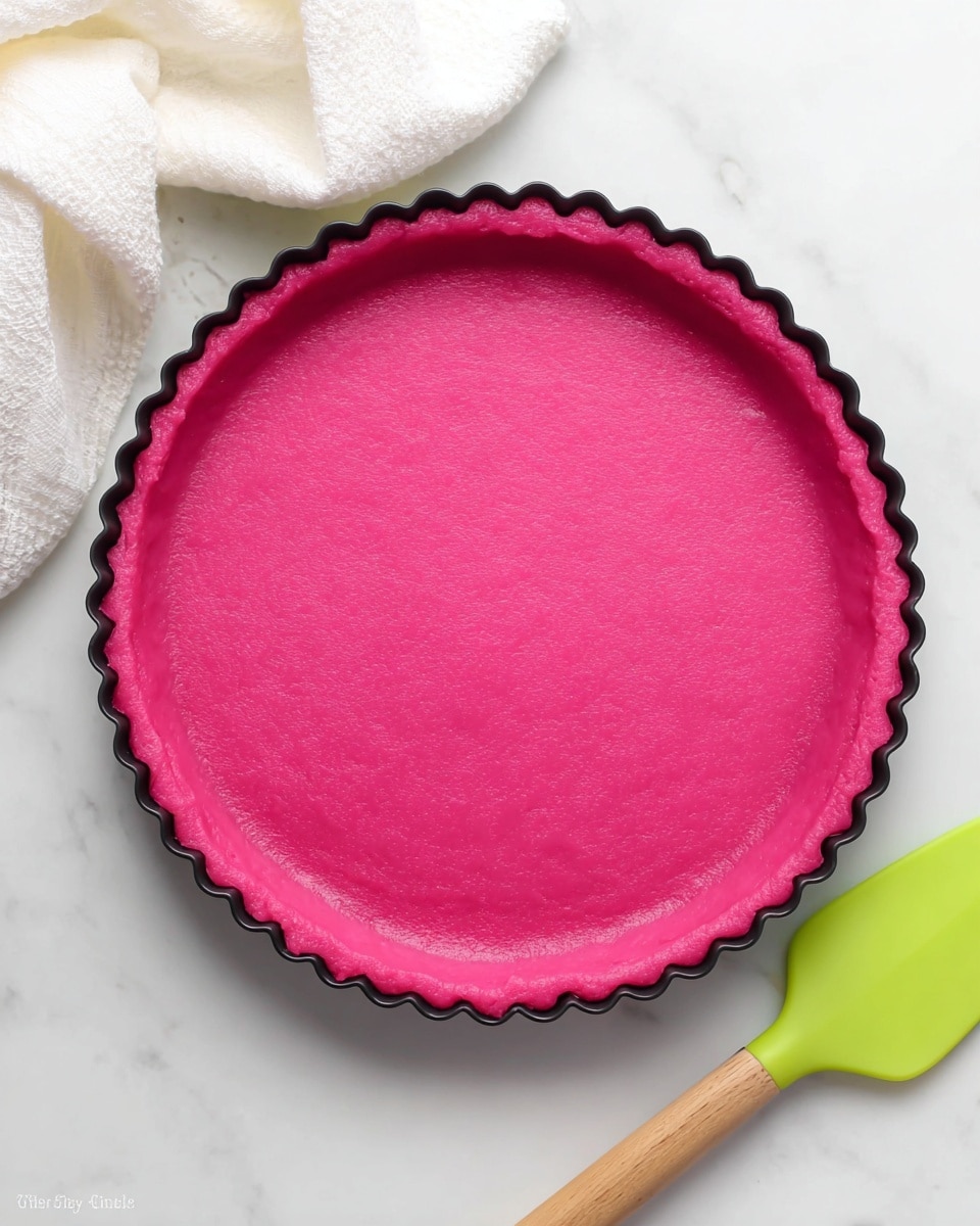 A single-layer round tart shell with scalloped edges is shown, filled with a smooth and thick bright pink dough pressed evenly along the bottom and sides. The tart shell sits in a black fluted tart pan placed on a white marbled surface, with a white cloth draped on the upper left corner. A lime green silicone spatula with a light wooden handle rests near the lower right edge of the tart pan. Photo taken with an iphone --ar 4:5 --v 7