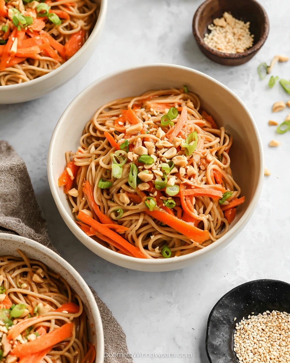 A large round bowl filled with light brown noodles mixed with thin orange carrot strips and small green onion slices on top, garnished with crushed peanuts scattered across; the bowl is placed on a white marbled surface with smaller white bowls containing similar noodle dishes, chopped peanuts, sesame seeds, and extra green onion slices around it. photo taken with an iphone --ar 4:5 --v 7
