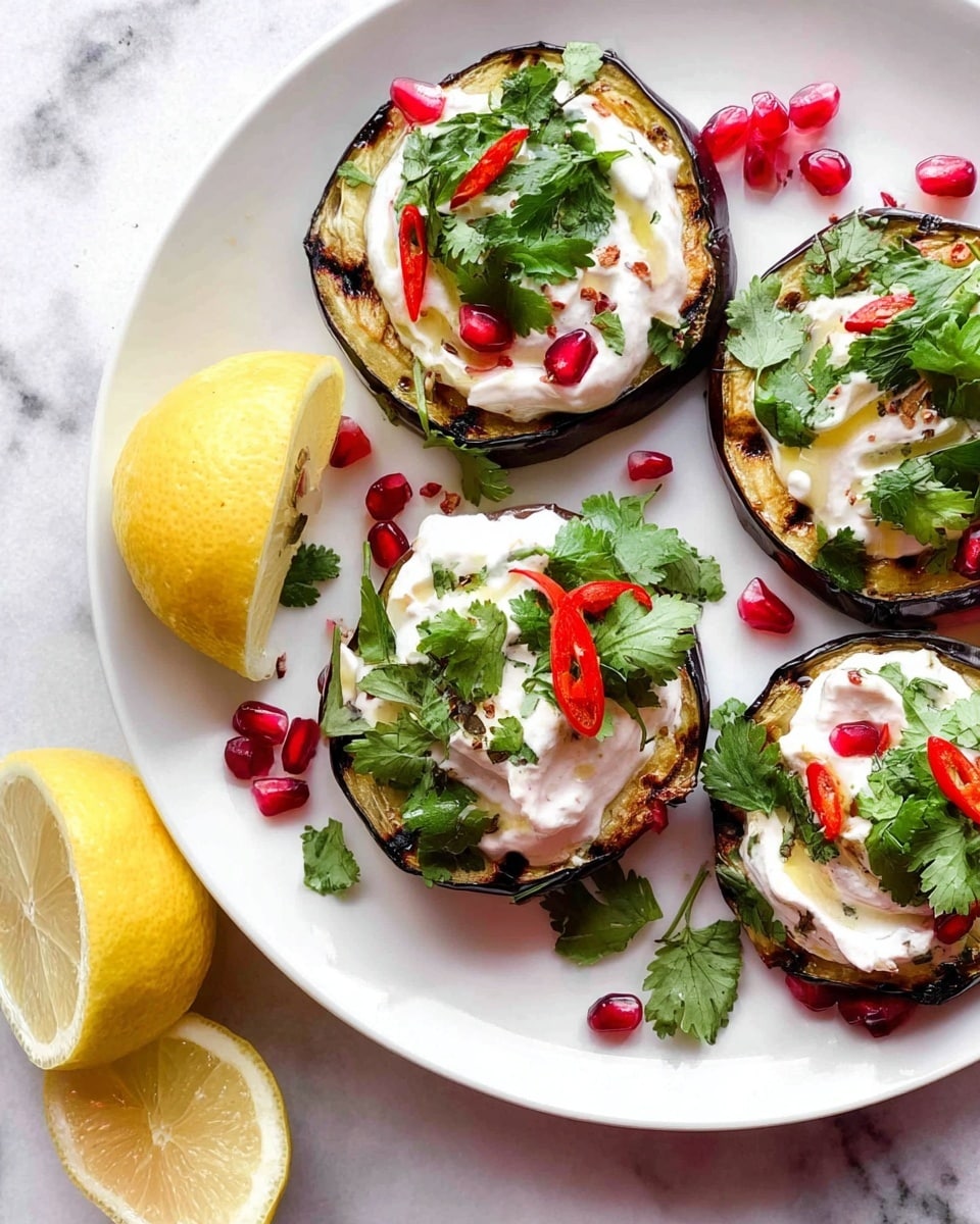 The image shows a white plate with four grilled rounds of eggplant, each topped with a thick layer of creamy white sauce. On top of the sauce, there are fresh green cilantro leaves, small slices of red chili peppers, and bright red pomegranate seeds scattered around, adding a pop of color. To the left side of the plate lies two halves of a yellow lemon, and the plate is placed on a white marbled surface. photo taken with an iphone --ar 4:5 --v 7