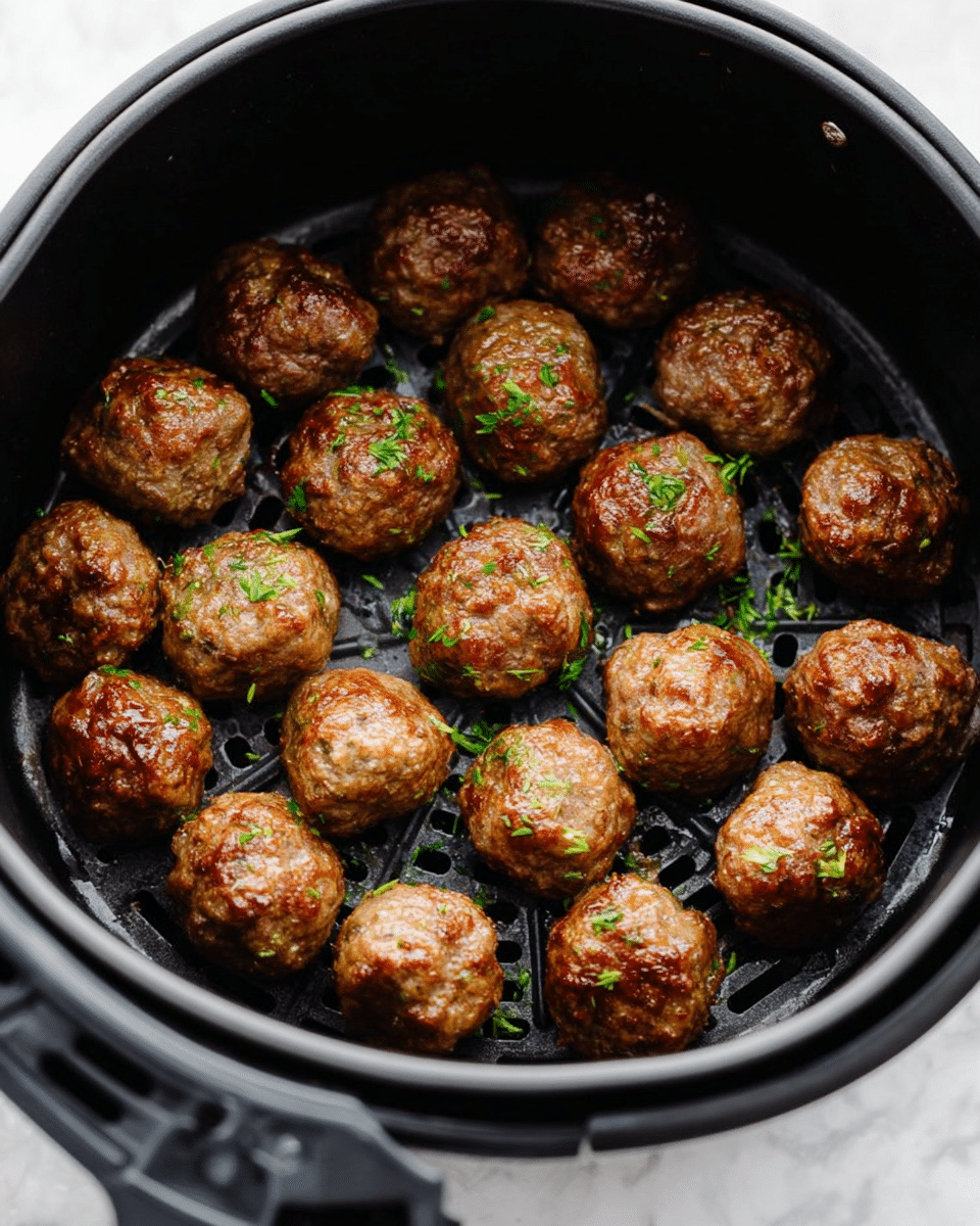 Inside a black air fryer basket, there are about 20 cooked round meatballs, arranged in a loose circle and some in the center. Each meatball is medium brown with a slightly shiny, crispy texture and has small green herb bits sprinkled on top. The basket’s surface is perforated with small holes, and the meatballs sit evenly spaced. The background below the air fryer is a white marbled texture. photo taken with an iphone --ar 4:5 --v 7