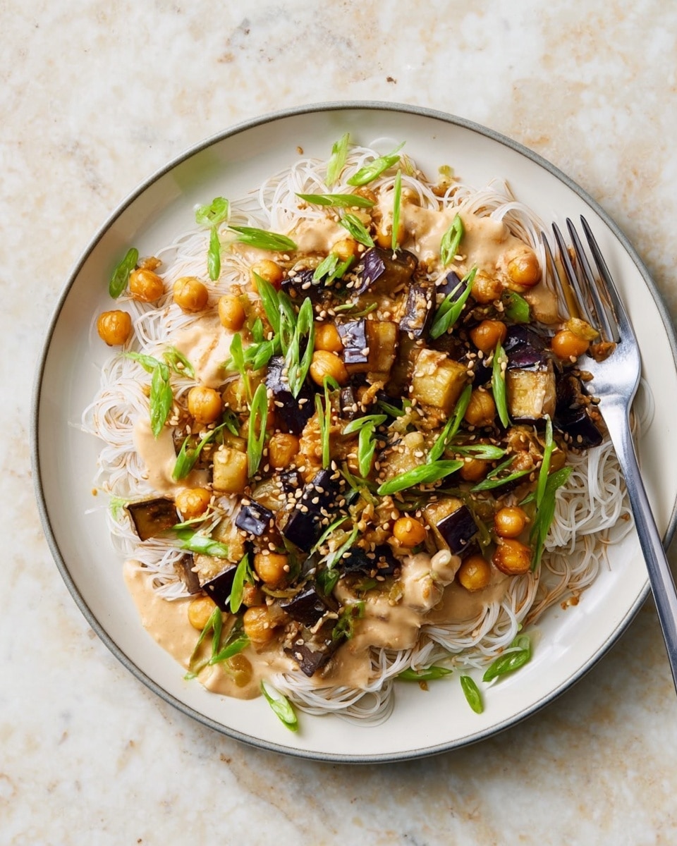 A plate of thin white rice noodles forms the bottom layer, topped with golden roasted chickpeas and browned eggplant chunks. Drizzled over everything is a creamy light brown sauce, and the dish is garnished with bright green sliced scallions and sprinkled with light tan sesame seeds. A silver fork lies on the right edge of the white plate, all placed on a white marbled surface. photo taken with an iphone --ar 4:5 --v 7