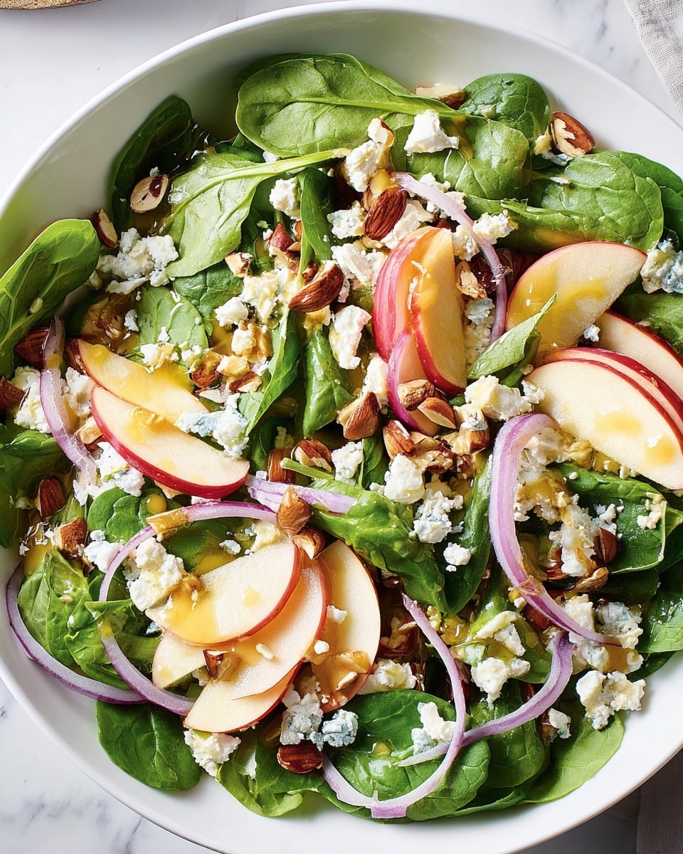 A white plate holds a fresh salad with bright green spinach leaves as the base layer, topped with thin slices of red apple with white flesh, and thin rings of purple red onion scattered throughout. Small white crumbles of cheese are sprinkled on top, alongside light brown sliced almonds. A light yellow dressing is drizzled over the salad, adding a glossy texture. A silver fork rests on the left edge of the plate, and the plate sits on a white marbled surface with a small glass container of dressing visible in the top left corner. Photo taken with an iphone --ar 4:5 --v 7