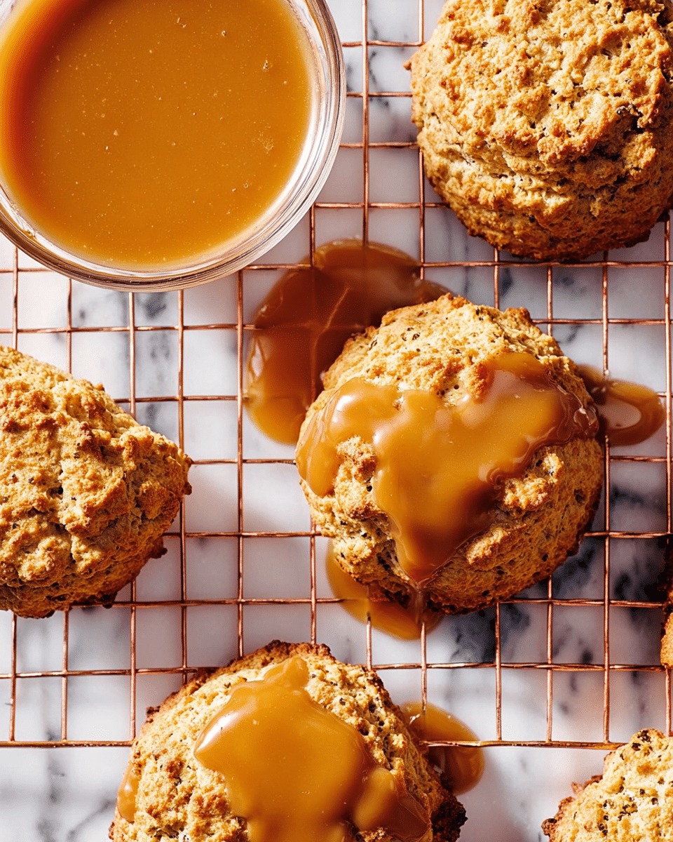 The image shows several round, golden-brown scones with a rough, crumbly texture placed on a copper wire cooling rack. Each scone has a thick and glossy caramel sauce dripping unevenly over the top, some pooling slightly on the wire rack below. To the top left, a clear glass bowl filled with more smooth, golden caramel sauce is visible. The background is a white marbled surface with soft shadows from the scones and rack. photo taken with an iphone --ar 4:5 --v 7