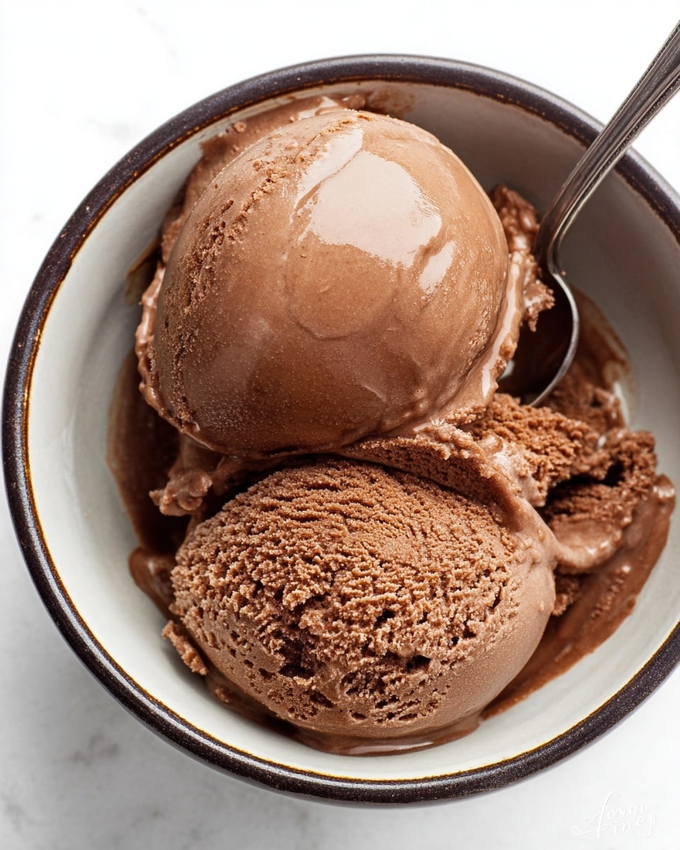 Two scoops of smooth, creamy chocolate ice cream are placed inside a white bowl with a dark rim. The top scoop has a shiny, soft texture with slight melting around the edges, while the bottom scoop shows a more granular, rich chocolate texture. A silver spoon is partially visible inside the bowl, resting against one side. The bowl is set on a white marbled surface. photo taken with an iphone --ar 4:5 --v 7