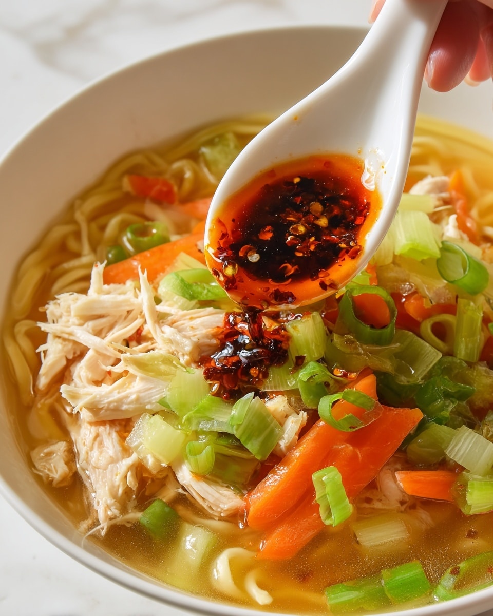 A close-up shot of a bowl of soup showing three main layers: at the bottom, light yellow noodles are partly visible; in the middle, bright orange carrot slices and green chopped celery mix together, with some light green onions scattered on top; above this, shredded white chicken pieces rest among the vegetables. A white spoon, held by a woman's hand, scoops dark red chili oil with visible chili flakes, spreading it over the soup's surface. The bowl is white and sits on a white marbled surface. photo taken with an iphone --ar 4:5 --v 7