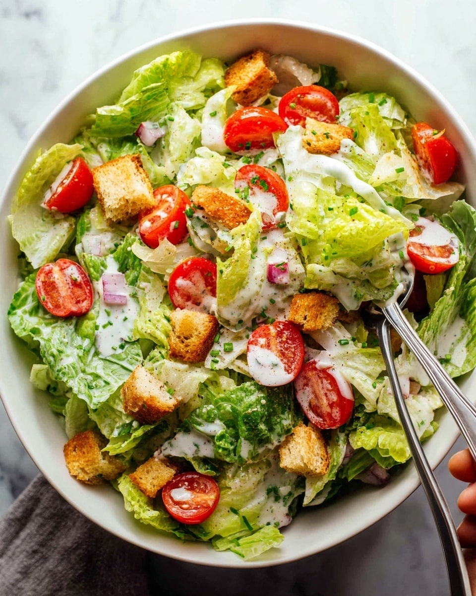 A large white bowl filled with layered fresh green lettuce leaves that have a crisp texture, topped with halved red cherry tomatoes scattered evenly across the salad. Small, golden brown croutons are sprinkled generously on top, adding crunch, while a creamy white dressing is drizzled over the salad with small pieces of finely chopped red onion and green chives spread throughout. Two silver forks rest on the edges of the bowl, one held by a woman's hand, dipping into the salad. The background is a white marbled surface that adds a clean, bright look to the scene, photo taken with an iphone --ar 4:5 --v 7