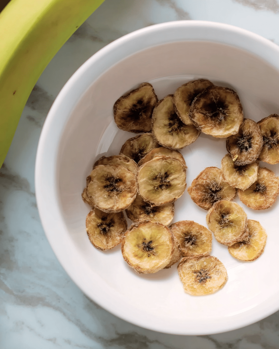 A white bowl contains a single layer of banana chips spread out loosely, each chip round with a shriveled texture. The chips have a light brown to dark brown gradient with darker spots in the middle, showing their dried, crispy nature. The bowl sits on a surface with a white marbled texture. Part of a green banana is visible in the top left corner. Photo taken with an iphone --ar 4:5 --v 7