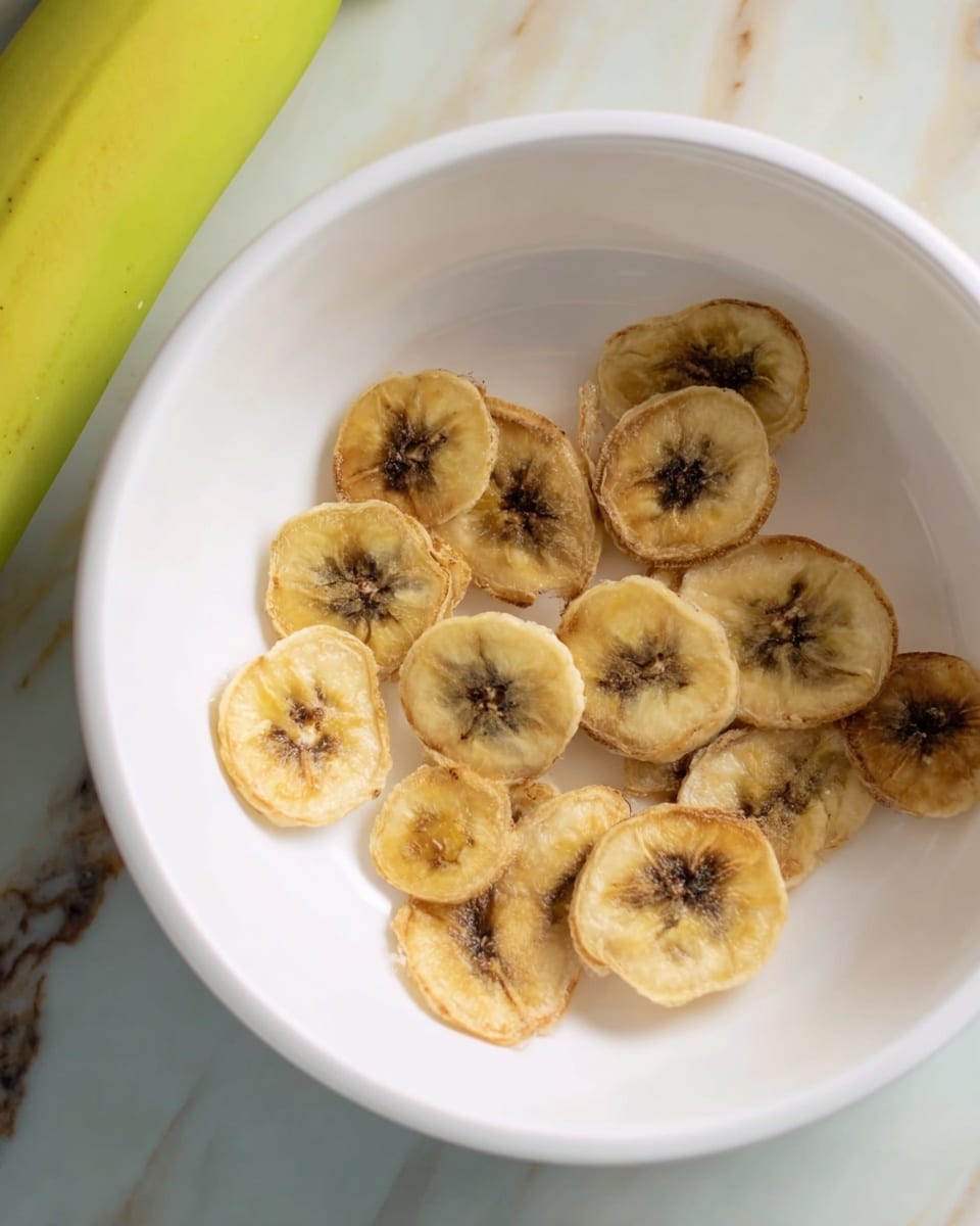 A white bowl contains about a dozen thin banana slices that are light yellow with dark brown spots and edges, showing signs of drying or crisping. The slices are spread inside the bowl with no stacking, and a part of a green banana is visible near the upper left side outside the bowl. The bowl sits on a surface with a white marbled texture. photo taken with an iphone --ar 4:5 --v 7