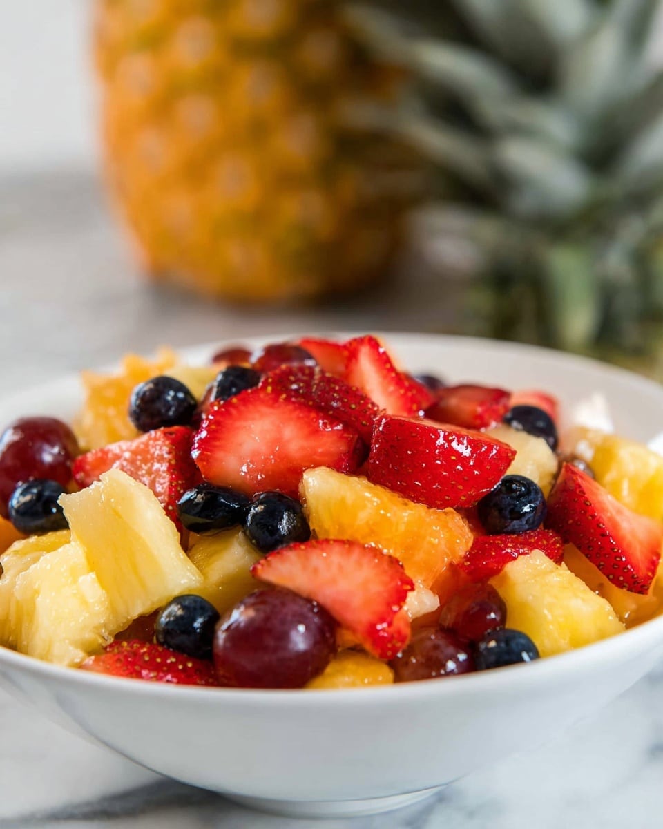 A white bowl filled with a colorful fruit salad showing at least four layers: the bottom layer has small orange-yellow pineapple chunks with a juicy texture, the next layer includes glossy red grapes and dark blueberries scattered throughout, above this are bright red sliced strawberries with a wet shine, and on top more mixed pieces of orange-yellow pineapple and red grapes, all sitting on a white marbled surface with a whole pineapple blurred in the background. photo taken with an iphone --ar 4:5 --v 7