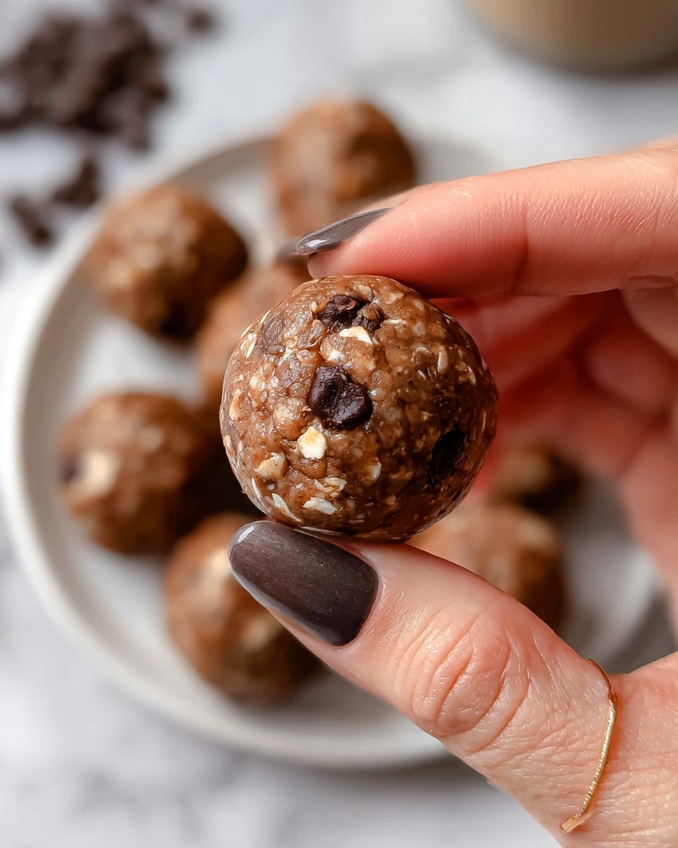 A close-up view of a round, brown energy ball with visible oat flakes and small dark chocolate chips mixed inside, held between a woman's thumb and fingers, showing natural skin tones and polished nails with a dark matte shade and a glossy light color, in front of a white plate filled with similar energy balls on a white marbled texture surface with scattered dark chocolate chips in the background, photo taken with an iphone --ar 4:5 --v 7