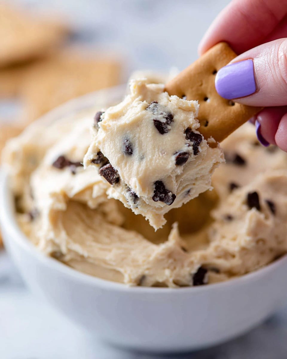 A close-up shows a white bowl filled with thick, creamy cookie dough that is pale beige with dark chocolate chips scattered throughout. A woman's hand with purple nail polish is holding a small rectangular cracker dipped in the dough, lifting it from the bowl. The dough clings to the cracker with visible chocolate chips on its surface. The background is a soft white marbled texture, out of focus. photo taken with an iphone --ar 4:5 --v 7