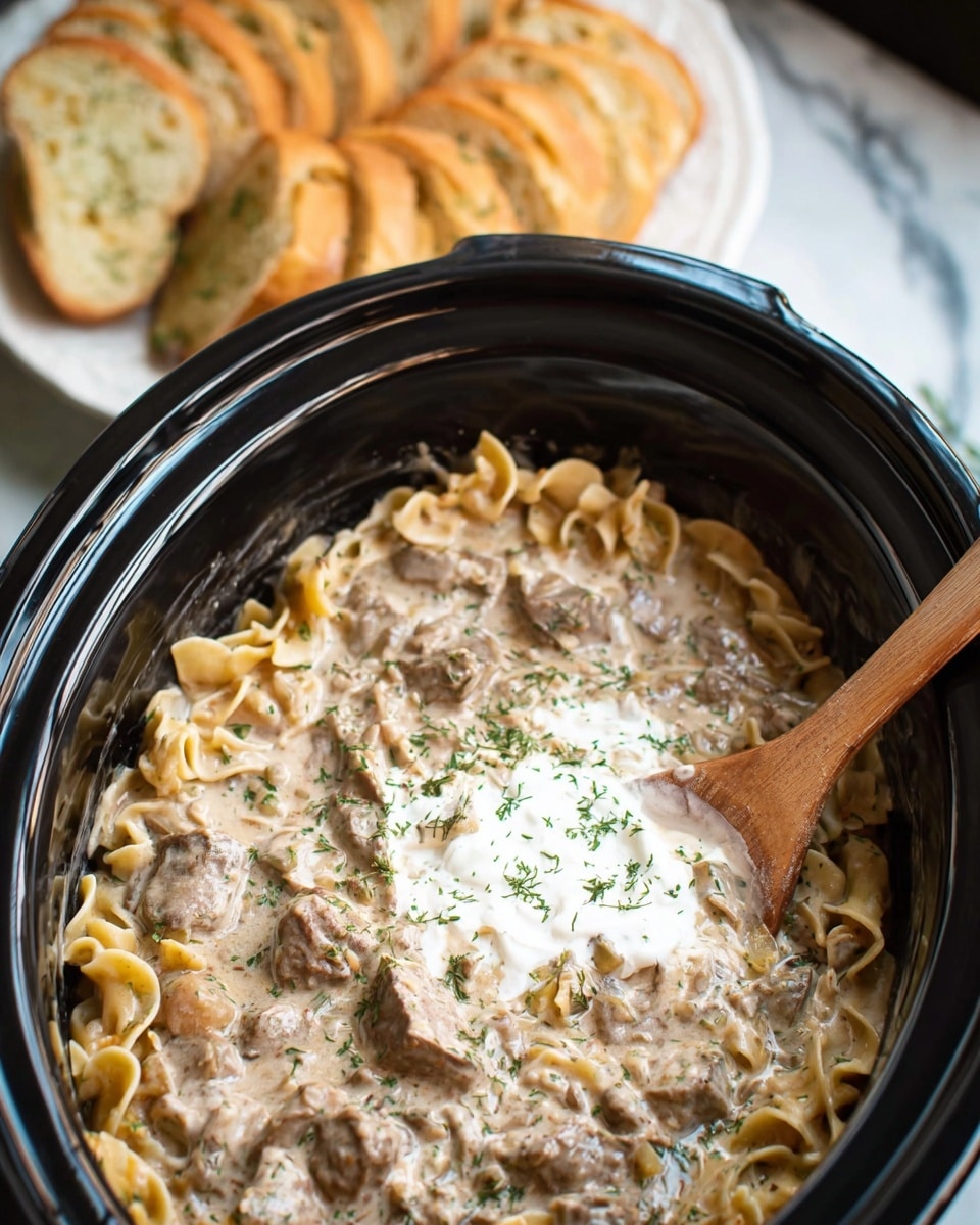 A large black slow cooker filled with creamy beef stroganoff, showing wide egg noodles mixed with bite-sized pieces of beef and mushrooms in a light brown, creamy sauce. A wooden spoon is stirring the dish, with some noodles and beef held above the surface. On the left side, a white plate holds slices of toasted garlic bread with green herb specks. Behind the plate, a white bowl with fresh green beans is visible. The slow cooker sits on a white marbled texture with a gray cloth nearby. Photo taken with an iphone --ar 4:5 --v 7