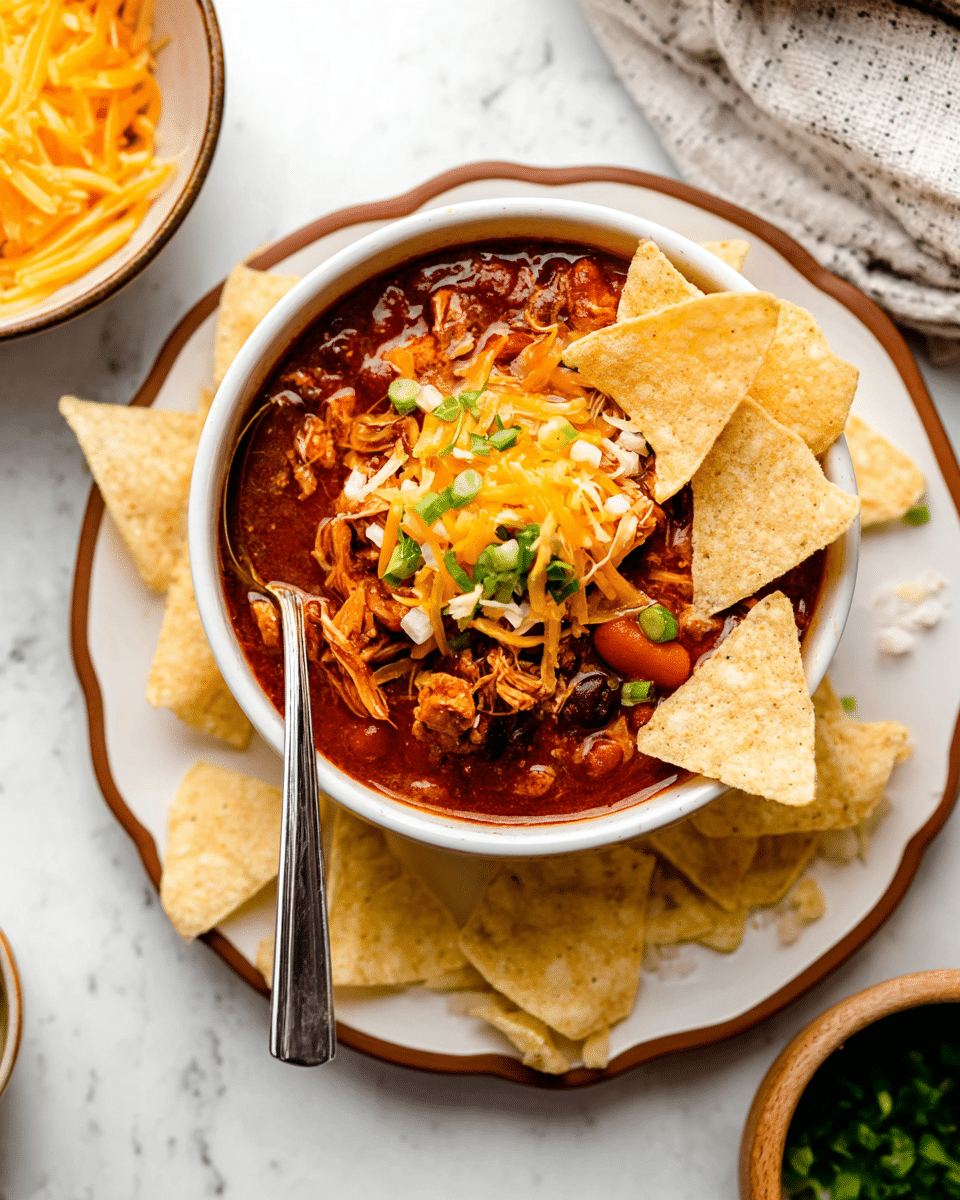 A white bowl filled with thick chili made of rich red sauce, beans, chunks of orange carrot, and shredded chicken is topped with bright orange shredded cheddar cheese and green chopped scallions scattered over the top. On the right side of the bowl, several triangular white tortilla chips stick into the chili with small chip crumbs on top. A shiny silver spoon is placed inside the bowl on the left, partially submerged in the chili. The bowl sits on a white plate with a brown scalloped edge and more white tortilla chips arranged around it. In the background, two bowls, one with more shredded cheddar cheese and the other with leafy greens, are partially visible over a white marbled textured surface. photo taken with an iphone --ar 4:5 --v 7