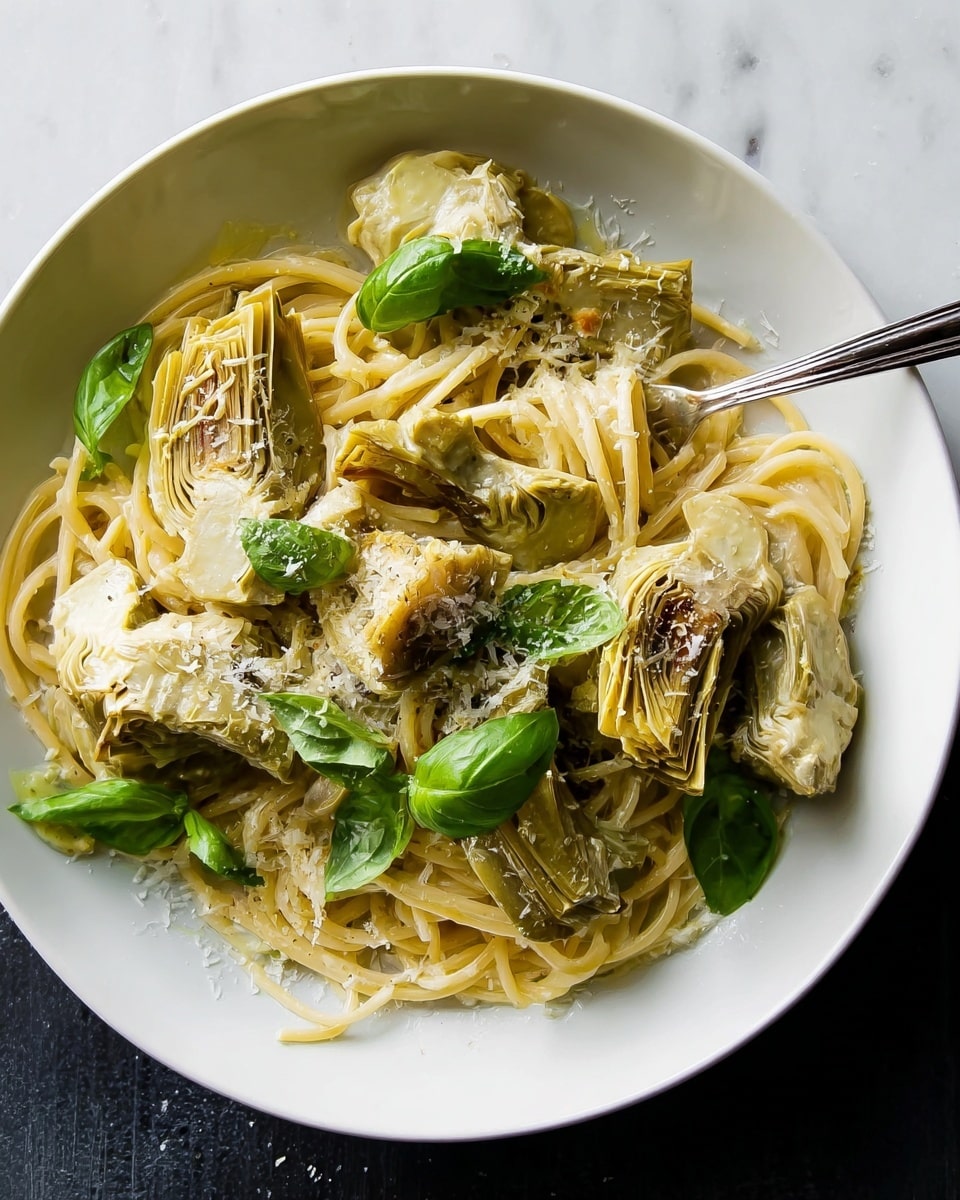 A white plate holds a serving of pasta with a creamy sauce, layered with pale yellow spaghetti noodles forming the base. On top, there are tender, light green artichoke pieces with some browned spots, showing a slightly rough texture. Scattered among the noodles and artichokes are fresh, bright green basil leaves that add a pop of color. The dish is lightly sprinkled with grated cheese giving a fine white powder effect. A silver fork is placed in the pasta, slightly buried under the noodles and leaves. The scene is set on a white marbled textured surface. photo taken with an iphone --ar 4:5 --v 7