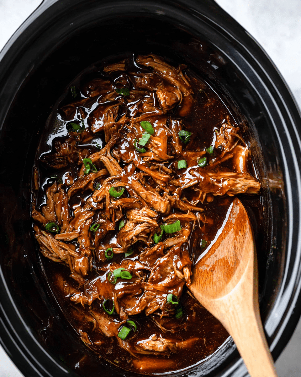 The image shows a close-up view of a slow cooker filled with cooked shredded chicken pieces covered in a shiny, dark brown sauce. The chicken is tender and moist, showing different sizes and shapes of shredded meat coated evenly in the sauce. Small pieces of chopped green onions are scattered on top, adding fresh green color contrast. A wooden spoon rests on the right side inside the cooker, partially submerged in the glaze. The slow cooker interior is black and glossy, and the whole scene sits on a white marbled surface. photo taken with an iphone --ar 4:5 --v 7