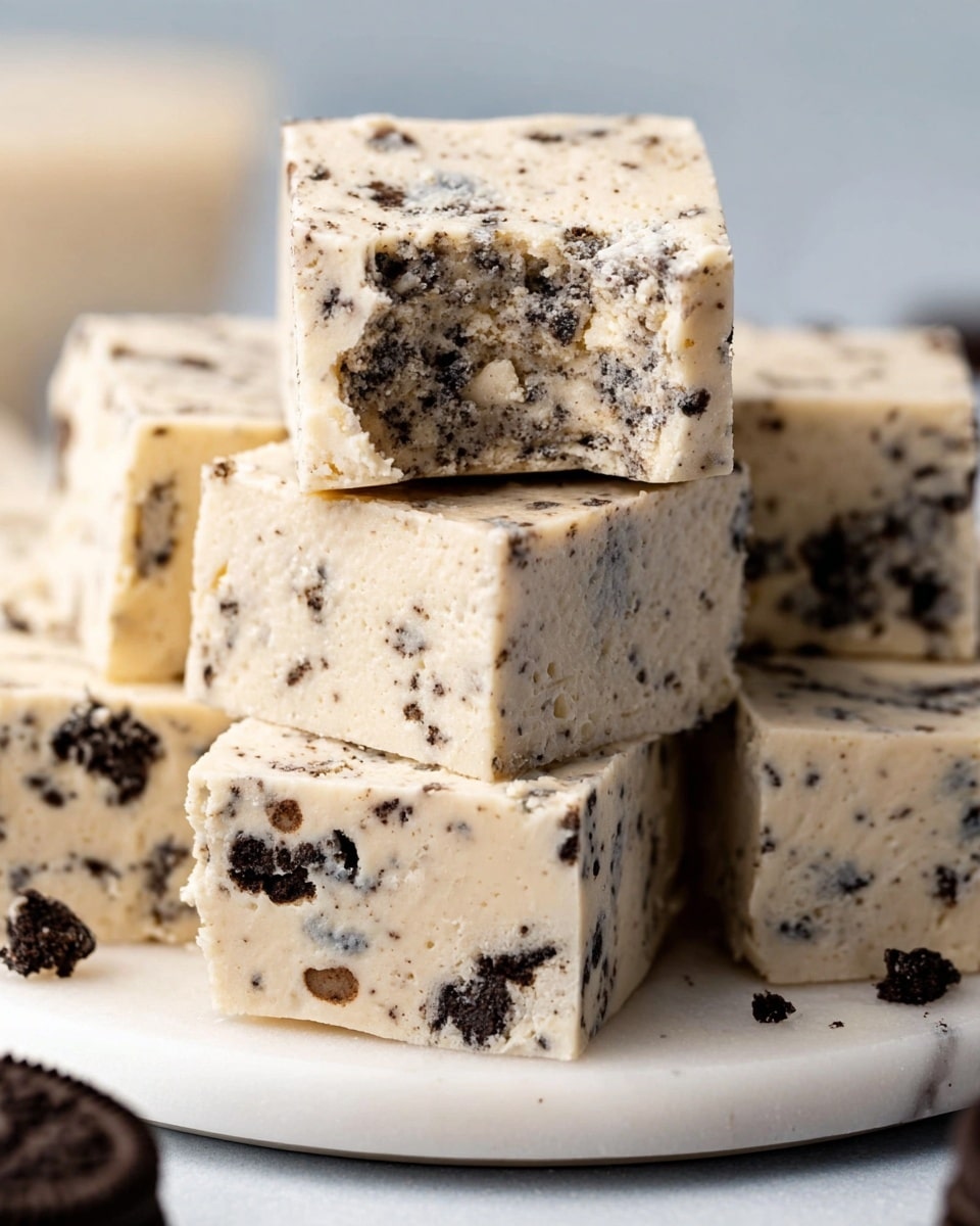 The image shows several square pieces of cookies and cream fudge stacked on a white plate, placed on a white marbled surface. Each piece has a creamy, off-white base mixed with small black cookie crumbs and chunks of white chocolate, giving it a speckled appearance. One piece is on top, slightly separated, with a small bite taken out, revealing a dense and smooth texture inside with visible cookie bits. The edges of the fudge are clean and squared, and there is a blurred Oreo cookie visible at the bottom of the plate. Photo taken with an iphone --ar 4:5 --v 7