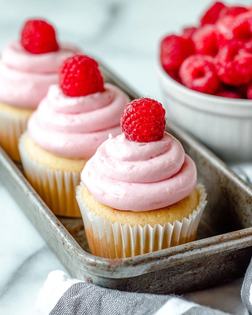 The image shows three cupcakes lined up on a rustic metal tray. Each cupcake has a light golden base with a white paper liner. On top of the base is a thick swirl of smooth, light pink frosting, forming three visible layers of soft curves. Each frosting swirl is topped with a single bright red raspberry that adds texture with its small seeds. To the right side of the tray, there is a white bowl partially filled with more red raspberries. The setting is on a white marbled surface, with a gray and white striped cloth partially visible near the tray. photo taken with an iphone --ar 4:5 --v 7