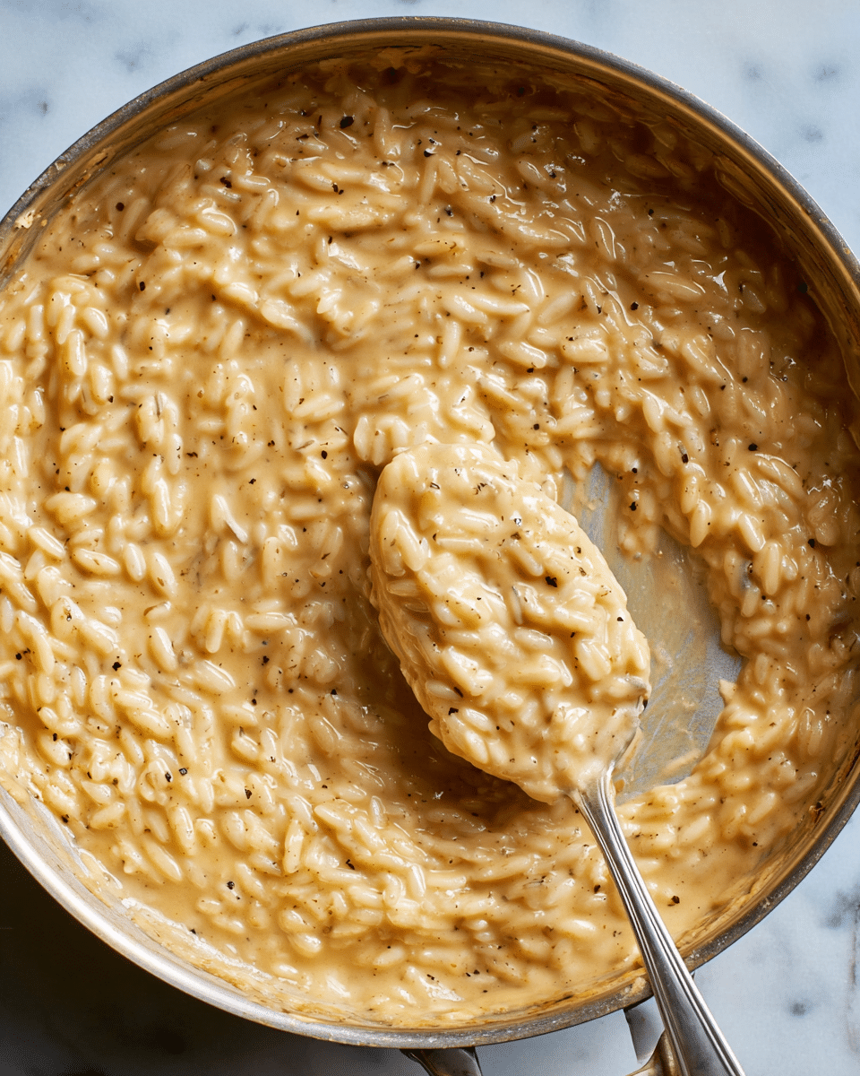 A close-up view of a large metal pan filled with creamy, light brown risotto with a thick texture and visible grains of rice coated evenly in the sauce. A metal spoon is scooping a portion from the center, showing the rice's slight stickiness and smooth creamy finish. The risotto has small black specks throughout, likely pepper or seasoning, and the pan edges show some glossy sauce residue. The pan rests on a white marbled texture surface. photo taken with an iphone --ar 4:5 --v 7