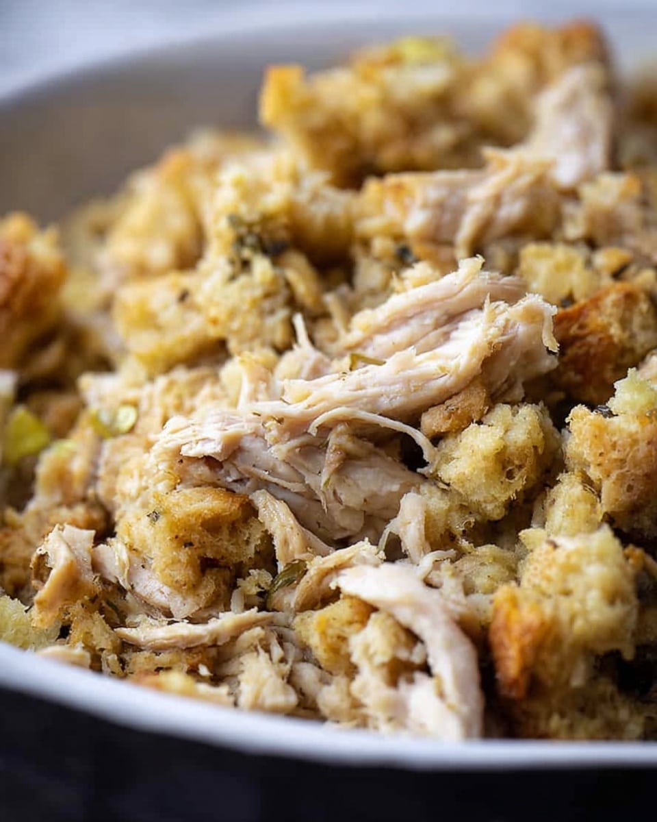 A close-up view of a dish made of shredded light beige chicken mixed with small, crumbly pieces of golden-brown stuffing. The texture looks soft and slightly moist with visible bits of herbs and spices throughout. It is served in a white bowl with a smooth edge, set against a white marbled texture in the background, giving focus on the detailed textures of the chicken and stuffing photo taken with an iphone --ar 4:5 --v 7