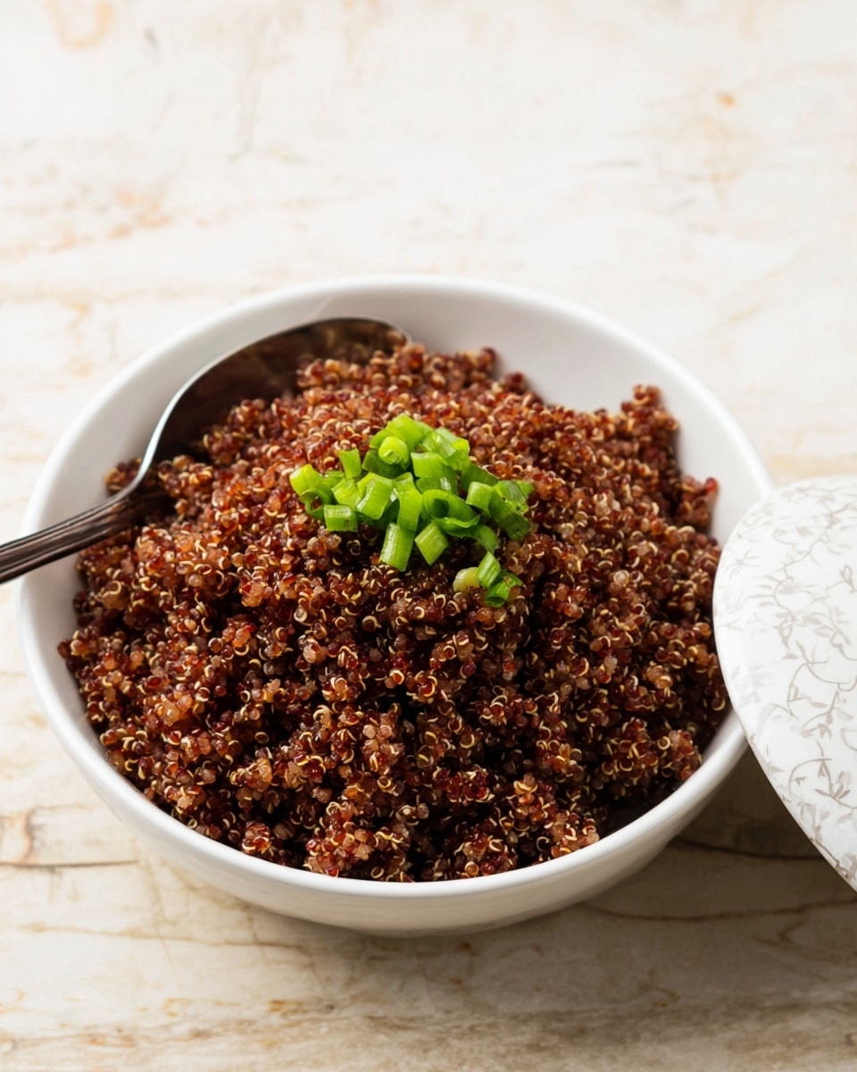 A white bowl filled with a mound of cooked red quinoa that has a textured, grainy appearance. The quinoa is topped with a small pile of bright green chopped scallions, adding a fresh contrast. A silver spoon rests partially inside the bowl on the left side, while on the right, a white patterned bowl lid leans against the bowl. The bowl is placed on a surface with a white marbled texture. Photo taken with an iphone --ar 4:5 --v 7