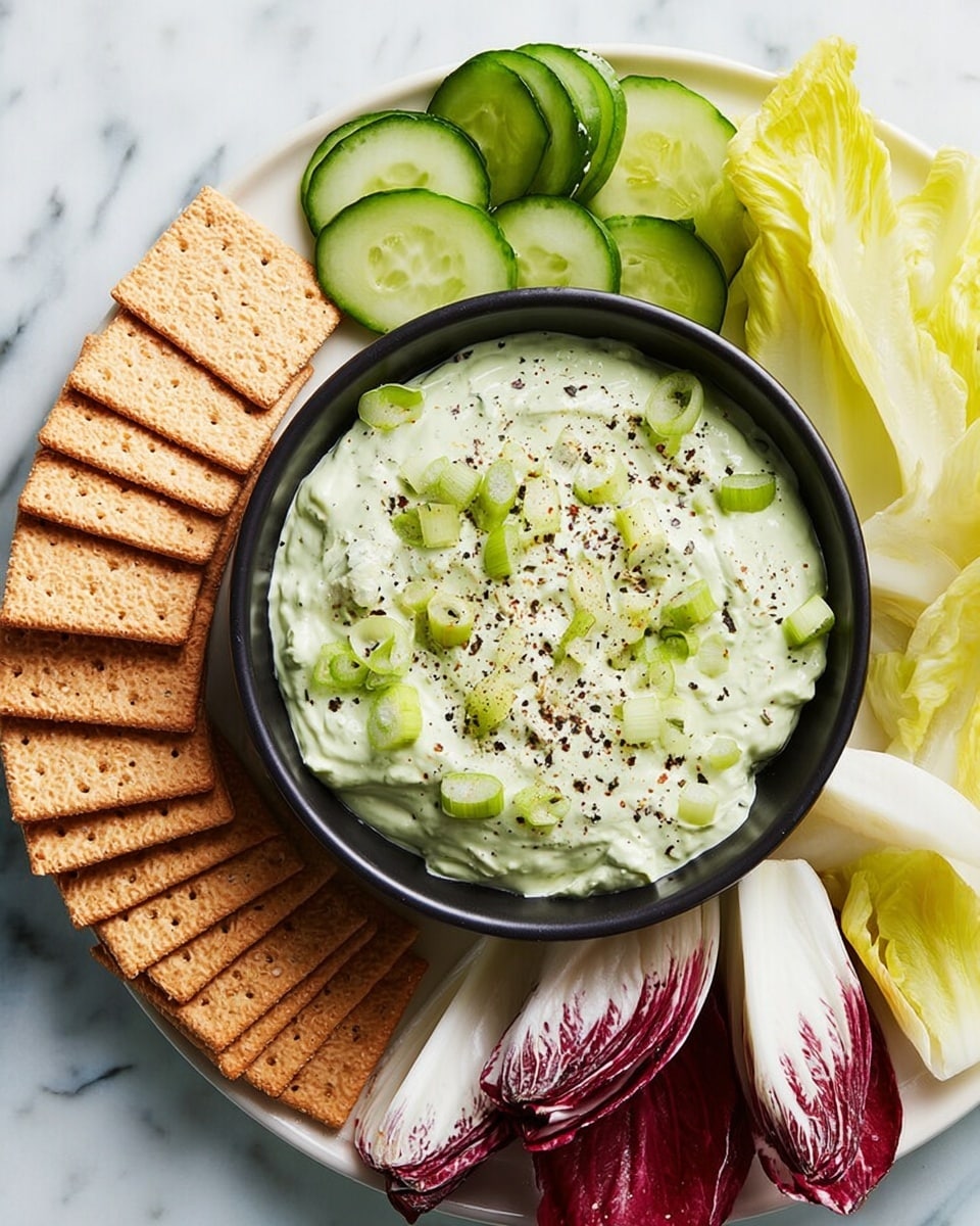 A white plate with a black bowl on top filled with green creamy dip sprinkled with small green onion pieces and black pepper on the surface; around the bowl, there are rectangular wheat crackers in a light brown color stacked in two layers, fresh cucumber slices that are shiny and green, thick white and deep red radicchio leaves layered loosely, and pale yellow-green chicory leaves placed near the crackers and cucumbers. The plate sits on a white marbled texture. photo taken with an iphone --ar 4:5 --v 7
