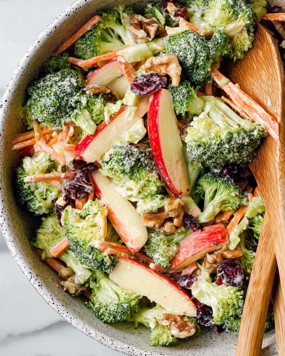 The image shows a fresh broccoli salad with several visible layers: bright green broccoli florets form the main layer, mixed with thin, orange shredded carrot strips scattered throughout; shiny red apple slices with a touch of cream dressing cover parts of the salad; pieces of light brown walnuts add texture in between; and small dark red dried cranberries are mixed all over. The salad is coated with a creamy, white dressing that lightly covers all ingredients. It is served in a white bowl with dark speckles, accompanied by two wooden spoons partially visible on the right side. The bowl sits on a white marbled surface. Photo taken with an iphone --ar 4:5 --v 7