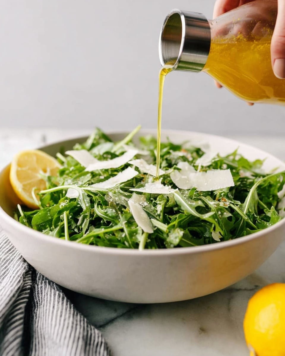 A large white bowl filled with fresh green arugula leaves and thin, light-colored shavings of cheese scattered on top. A bright yellow lemon wedge sits on the side of the greens, adding a pop of color. Above the bowl, a woman's hand is holding a metal-top glass bottle, pouring a golden dressing over the salad. The bowl is set on a white marbled surface with a soft focus gray and white striped cloth in the background. Photo taken with an iphone --ar 4:5 --v 7