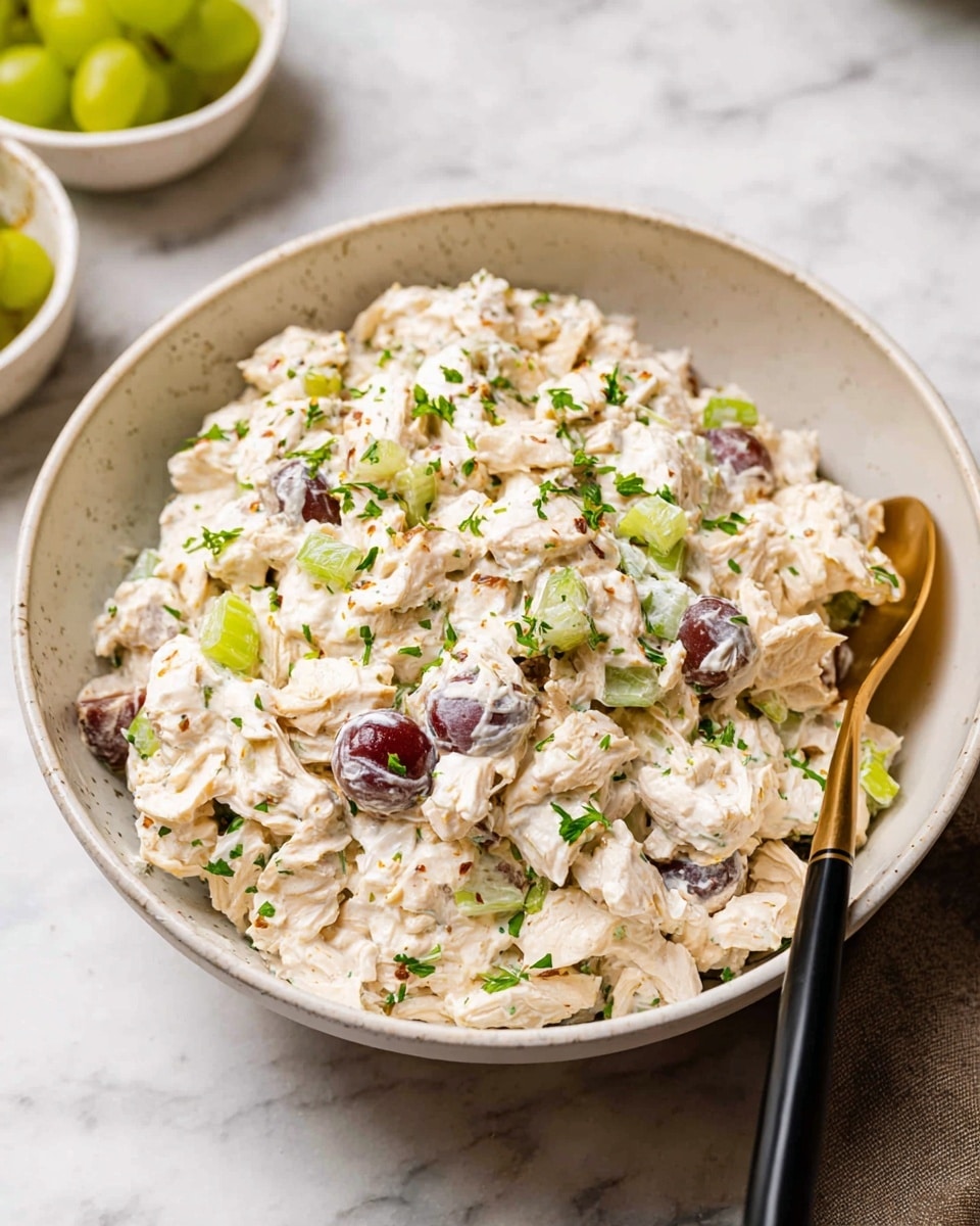 A round white bowl filled with creamy chicken salad containing visible chunks of white chicken, grape halves, small pieces of green celery, and fresh green herbs sprinkled on top, all mixed in with a light, thick white dressing; a black and gold spoon is partially inserted on the right side of the bowl, which sits on a white marbled surface with two small white bowls in the background holding grapes and celery pieces, photo taken with an iphone --ar 4:5 --v 7