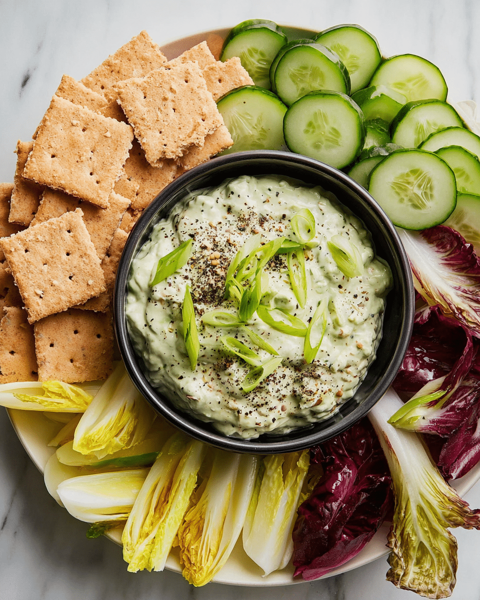 A close-up view of a white plate with several layers of food arranged around a black bowl in the center. The top layer of the plate has light brown square crackers stacked in a pile on the left and right sides. Below and around the crackers are slices of fresh green cucumber, some with dark green skins, overlapping each other. Mixed with the cucumber slices are leaves of pale yellow and purple-red endive, creating a colorful, layered border. In the black bowl in the middle is a creamy, light green dip, textured with small bits and topped with sliced fresh green onions and a sprinkle of coarse black pepper. The whole dish is set on a white marbled texture. photo taken with an iphone --ar 4:5 --v 7