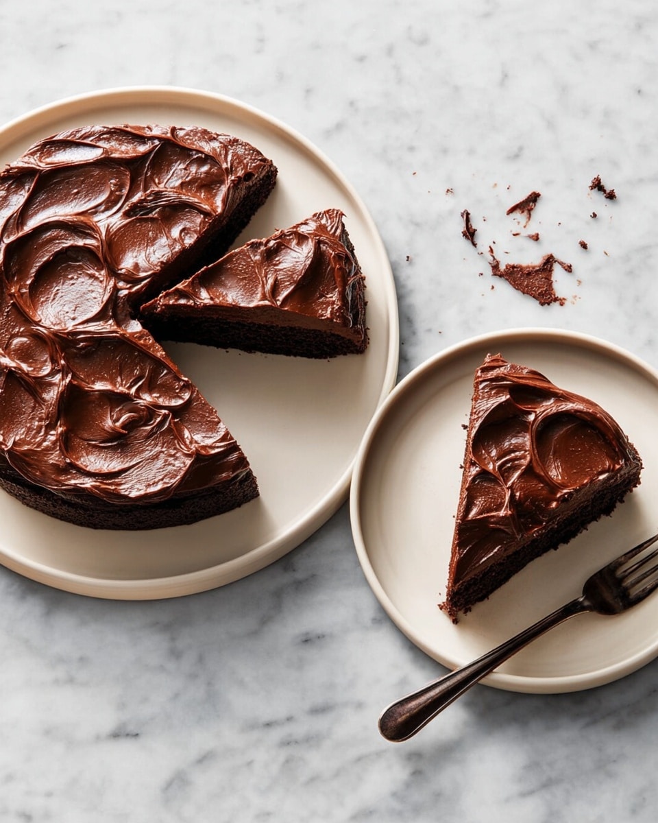 A round chocolate cake with one slice cut out and placed on a smaller white plate beside it. The cake has one thick dark brown base layer topped with a glossy, smooth and swirled dark chocolate frosting that looks creamy and rich. Both plates sit on a white marbled surface. Next to the slice on the smaller plate is a dark bronze fork. There are a few crumbs scattered around the plates. Photo taken with an iphone --ar 4:5 --v 7