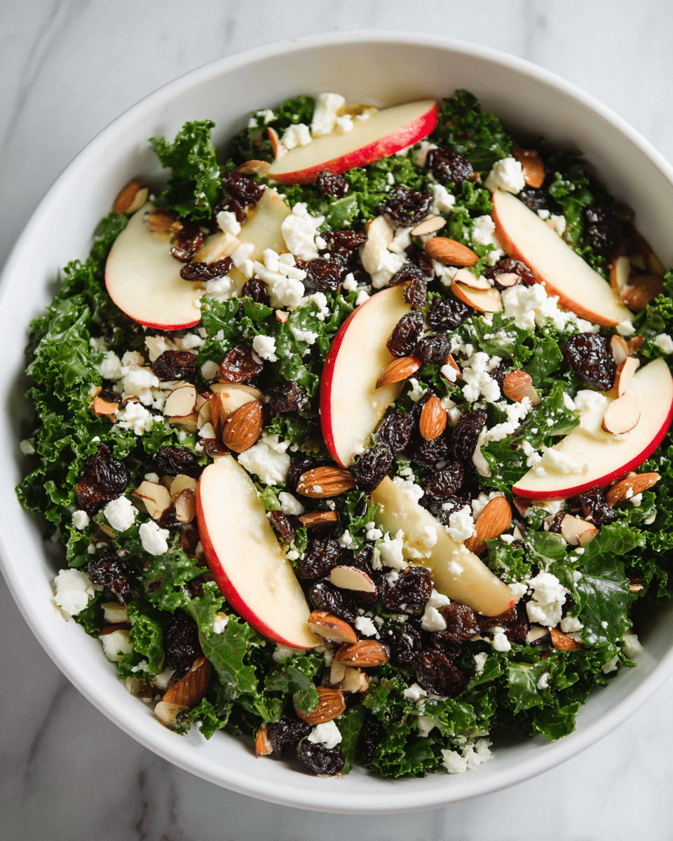 A white bowl filled with a fresh kale salad that has ruffled dark green leaves as the base layer. On top, there are thin slices of red-skinned apple with a smooth texture, juicy dark raisins scattered unevenly, and small crunchy light brown almond slices sprinkled all over. Crumbled white cheese pieces are spread across the salad, adding contrast to the vibrant colors. The bowl is placed on a white marbled surface. photo taken with an iphone --ar 4:5 --v 7
