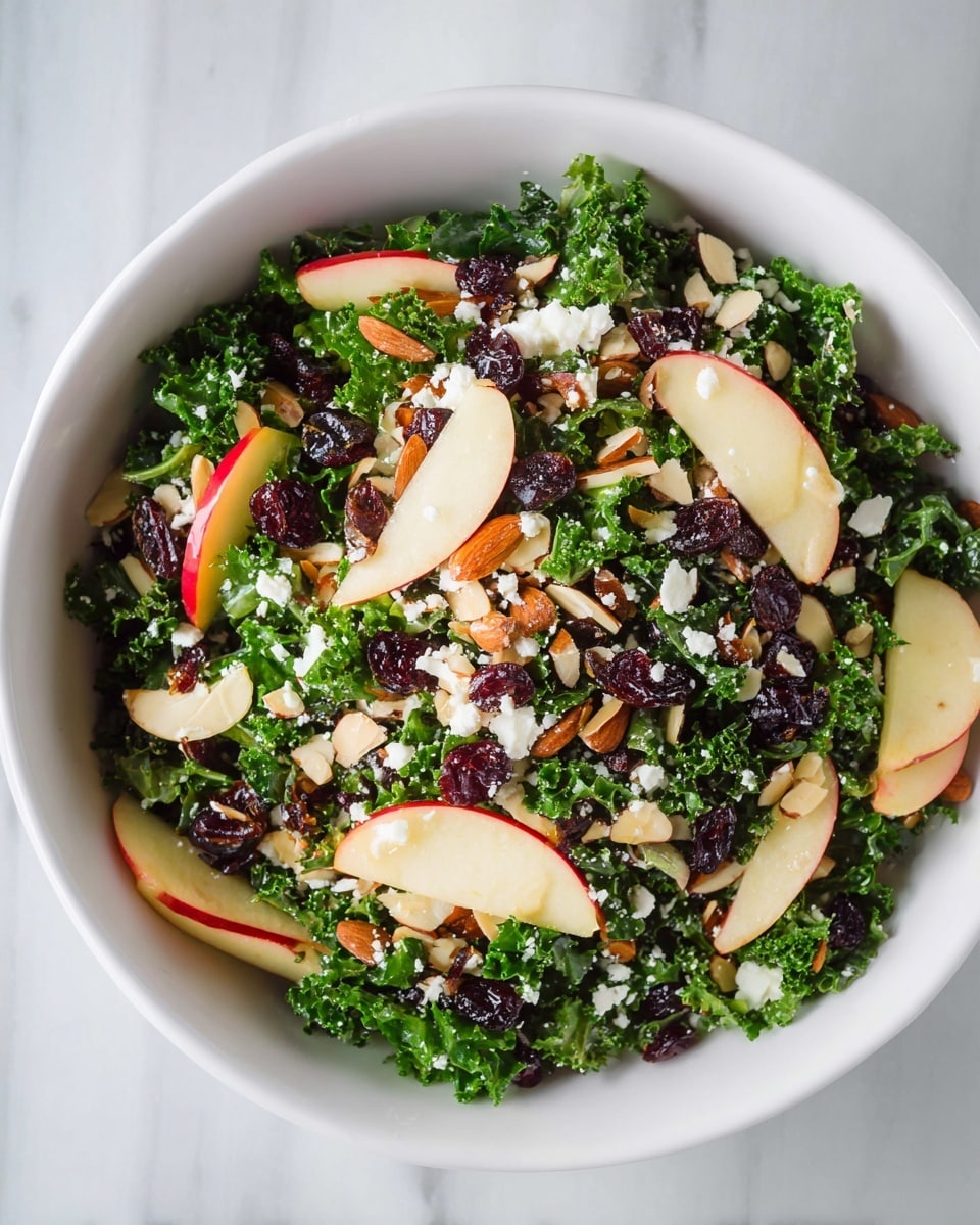 A white bowl filled with a green kale salad as the base layer, topped with thin apple slices showing red and white colors, dark dried cranberries scattered evenly, light brown toasted almond slices spread throughout, and small white crumbles of cheese sprinkled on top, all set on a white marbled surface photo taken with an iphone --ar 4:5 --v 7