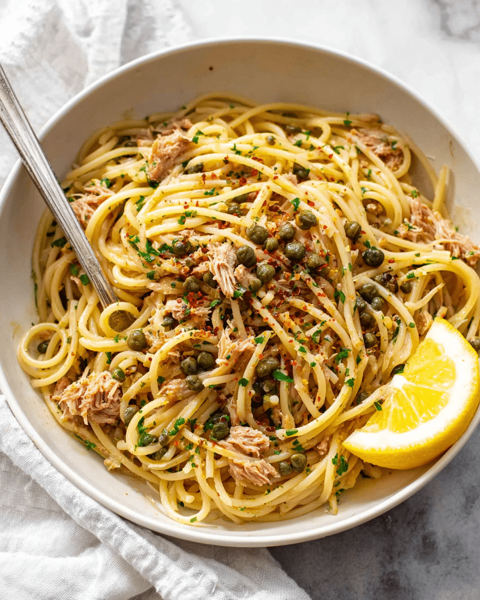A bowl of spaghetti pasta mixed with pieces of tuna and green capers, topped with small bits of chopped parsley and sprinkled with red pepper flakes, creating a mix of brown, green, and yellow colors. The pasta is light golden and glossy, tangled in the center of a white bowl. A silver fork is resting on the side of the bowl, and a yellow lemon wedge is placed to the right, adding color contrast. The bowl sits on a white marbled surface with a white cloth underneath. Photo taken with an iphone --ar 4:5 --v 7