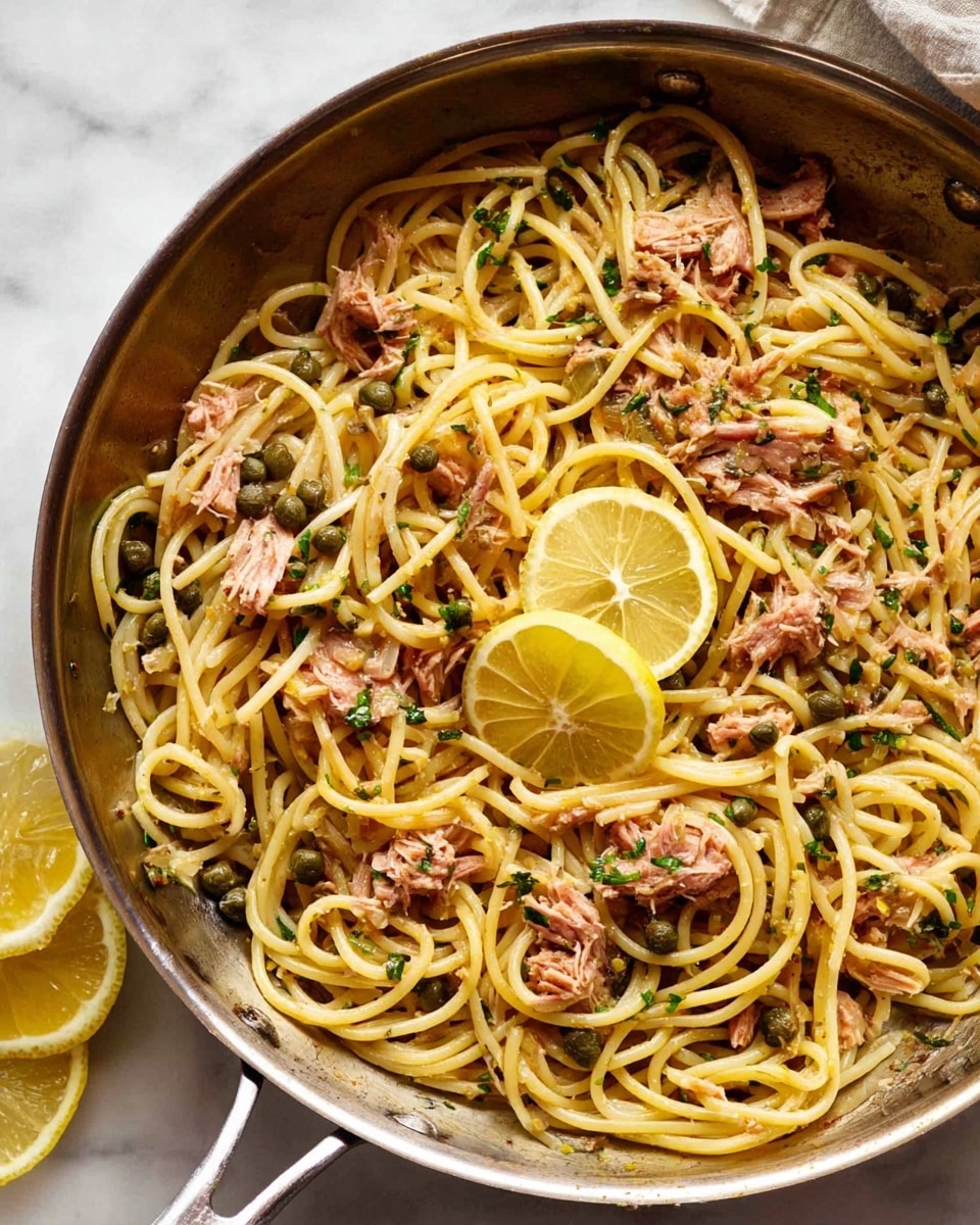 A metal pan filled with spaghetti pasta layered with small chunks of light pink tuna, scattered green capers, and bits of chopped herbs. On top, two thin, translucent lemon slices rest near the center, adding a subtle yellow touch. The background shows a white marbled surface with a few lemon slices off to the side. The pasta looks glossy as if lightly coated in oil, and the colors are warm and natural. photo taken with an iphone --ar 4:5 --v 7