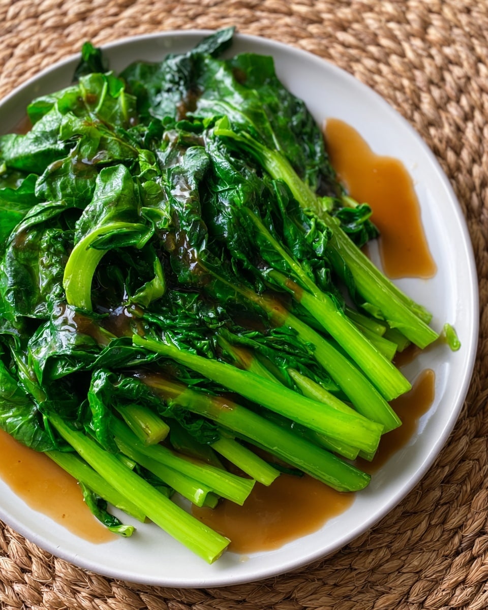 A white plate holds a simple dish of bright green steamed or boiled leafy greens with long stems and dark green leaves, covered in a glossy, light brown sauce. The stems are stacked in a slightly messy pile on the plate, with some leaves spread flat on top. The sauce pools slightly around the base of the greens. The plate is placed on a woven, natural textured mat, adding a warm, rustic contrast to the fresh greens and white plate. Photo taken with an iphone --ar 4:5 --v 7