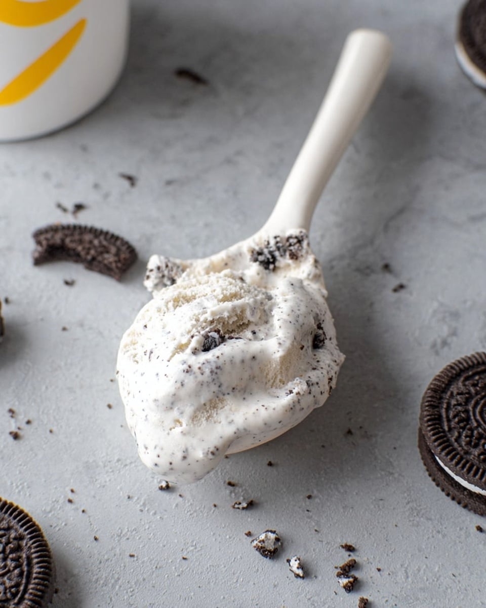 A close-up image of a white ceramic spoon filled with cookies and cream ice cream, showing a creamy white base with small dark cookie bits mixed throughout. The spoon rests on a light gray stone surface with a few scattered broken pieces of chocolate sandwich cookies around it. The top part of a white cup with a yellow design is faintly visible in the top left corner. The texture of the ice cream is smooth with visible cookie chunks, and the scene has a clean, simple look. Photo taken with an iphone --ar 4:5 --v 7
