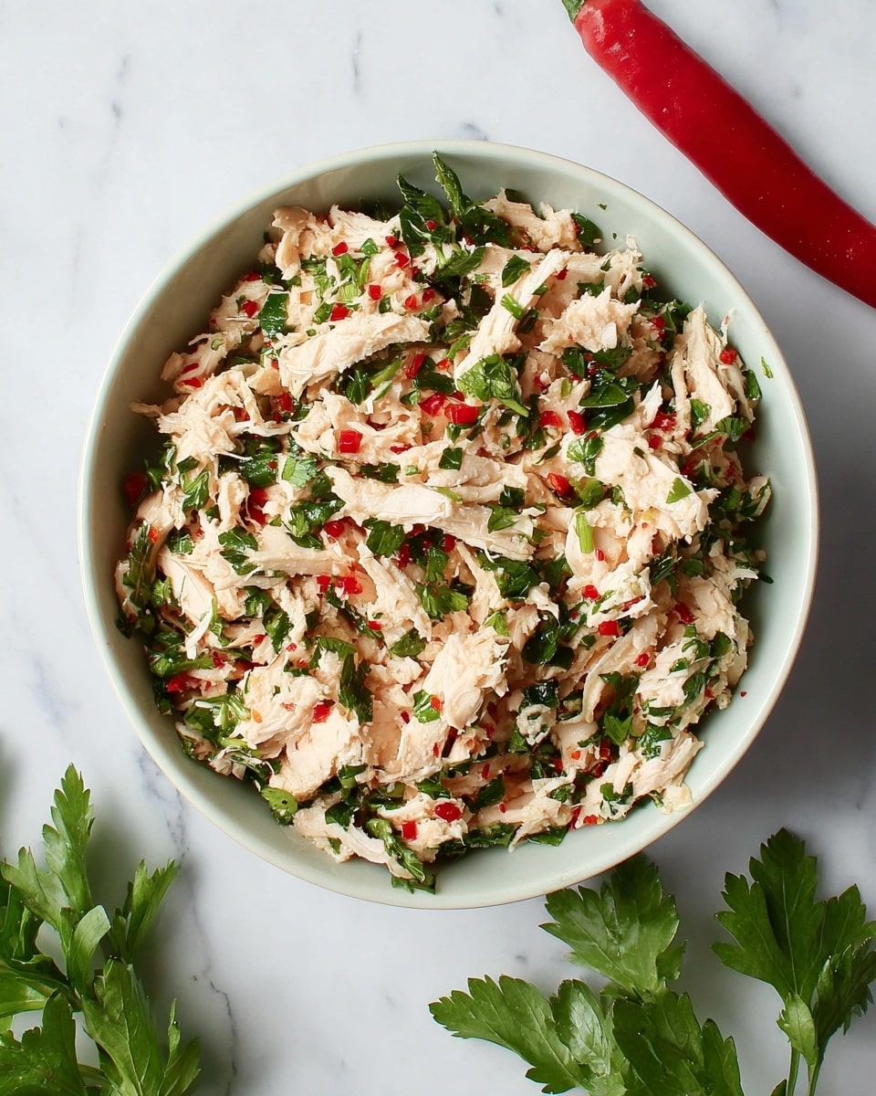 A white bowl filled with a mix of shredded pale pink cooked chicken and finely chopped vibrant green herbs, scattered with small pieces of bright red chili peppers throughout, all sitting on a white marbled surface, with a whole red chili pepper to the top right and fresh green parsley leaves to the bottom left, photo taken with an iphone --ar 4:5 --v 7