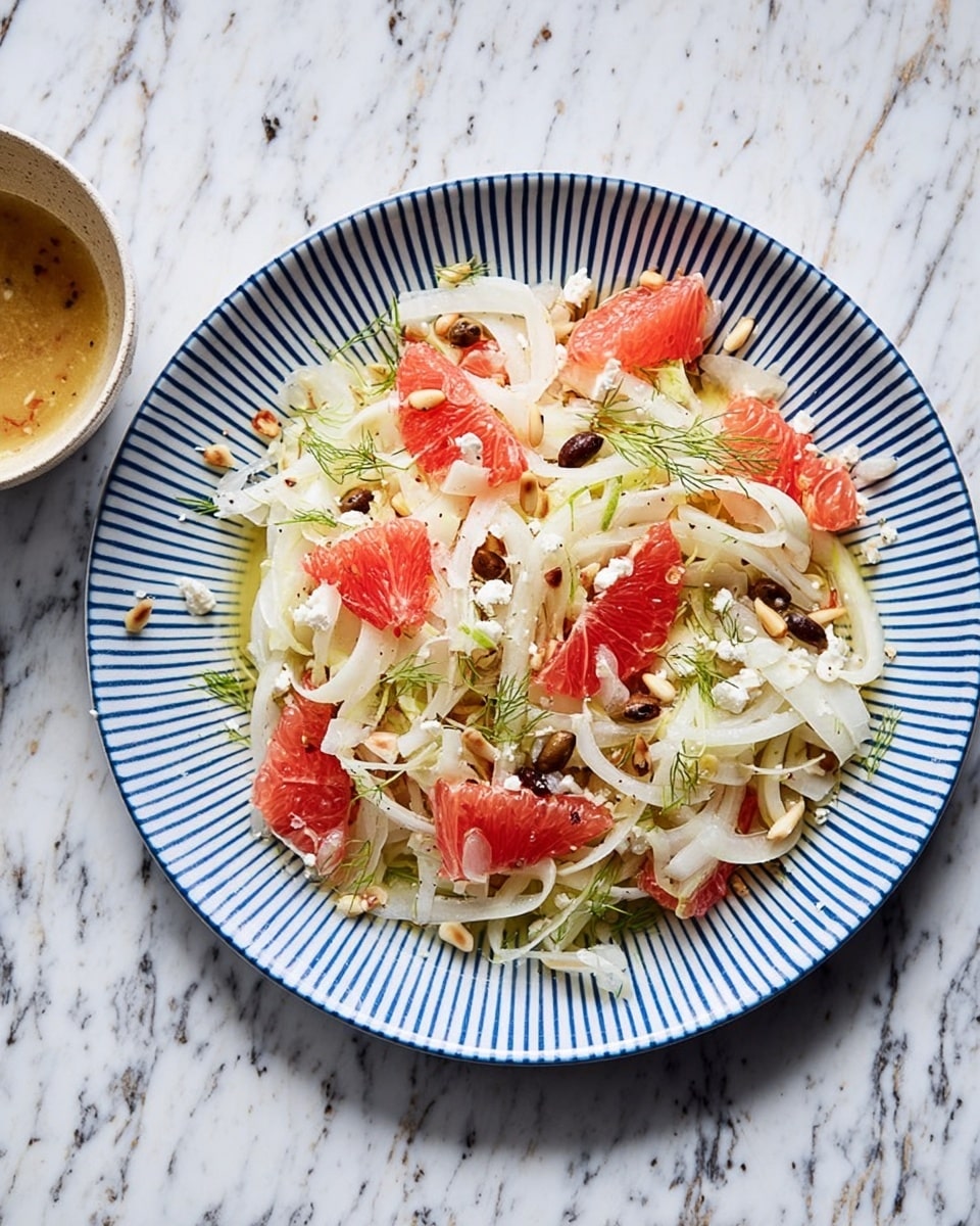 The image shows a round white plate with blue stripes filled with a light and fresh salad. The bottom layer is thin, white, curly slices of fennel forming a tangled base. On top of that, there are bright pink grapefruit segments scattered evenly, adding vibrant color. Small dark brown olive slices and crumbly white cheese bits are sprinkled over the salad. Tiny toasted pine nuts add a touch of golden brown and texture. Delicate green fennel fronds are placed lightly as garnish. The plate is set on a white marbled surface with a small bowl of light brown dressing with herbs seen in the corner. photo taken with an iphone --ar 4:5 --v 7