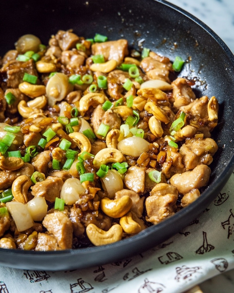 A close-up view of a black pan filled with cooked chicken pieces, light brown in color with a slightly glossy texture from sauce, mixed with whole cashew nuts that are golden brown and shiny. There are sliced water chestnuts that are pale beige and firm-looking, scattered throughout the pan. The dish is garnished with small pieces of bright green chopped green onions, adding a fresh pop of color on top. The pan sits on a white marbled surface with a glimpse of a cloth with a simple line drawing. Photo taken with an iphone --ar 4:5 --v 7