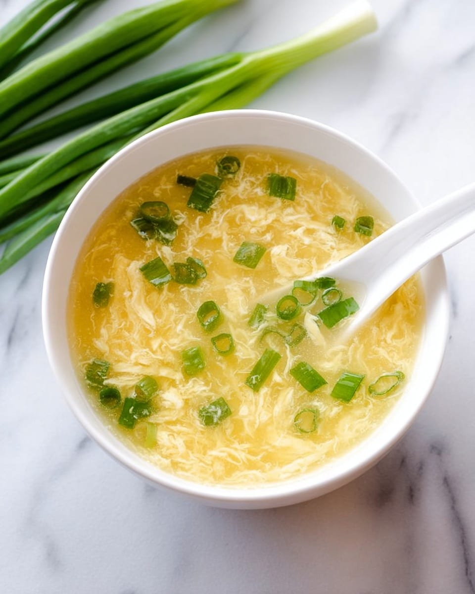 A white bowl filled with light yellow egg drop soup with soft, thin swirling egg ribbons creating a delicate texture throughout; scattered on top are bright green chopped scallions adding a fresh color contrast. A white ceramic spoon is resting inside the bowl, partly submerged in the soup. In the background, fresh green scallion stalks lie against a white marbled surface. photo taken with an iphone --ar 4:5 --v 7