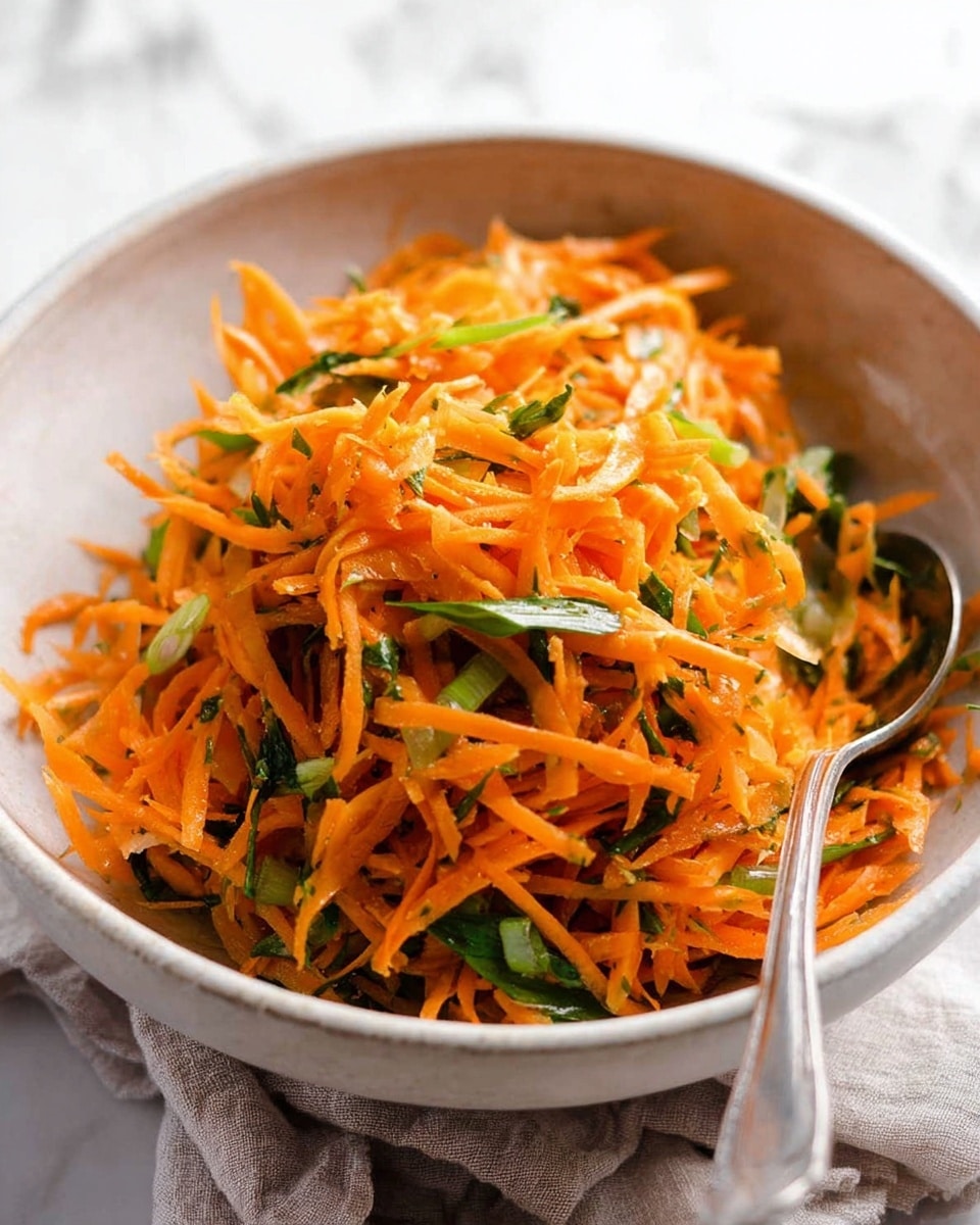 A close-up of a bowl filled with shredded carrot salad mixed with thin green vegetable slices, likely green onions, creating a layered mix of bright orange and fresh green colors. The carrot strands are piled loosely on top of each other, with the green pieces scattered evenly throughout, adding texture and contrast. The bowl is white and has a soft round shape with a slightly rough texture. A silver spoon is placed inside the bowl, partially buried in the salad. The bowl sits on a white marbled surface with a cloth cloth nearby. Photo taken with an iphone --ar 4:5 --v 7