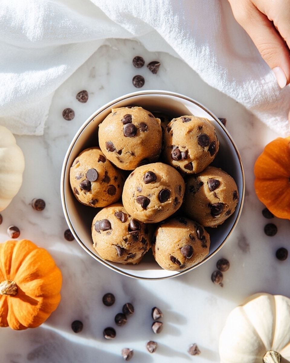 A white bowl filled with about ten round cookie dough balls studded with dark chocolate chips, each ball showing a smooth, slightly cracked light brown dough with spots of shiny, rich chocolate. The bowl is placed on a white marbled surface scattered with loose chocolate chips, surrounded by two small whole orange pumpkins and one white pumpkin, adding bright pops of color to the scene. A white cloth is partly visible in the corner next to the bowl, and a woman's hand reaches gently toward the bowl from the left side. The light is bright and natural, highlighting the soft texture of the dough and the smooth marbled surface photo taken with an iphone --ar 4:5 --v 7