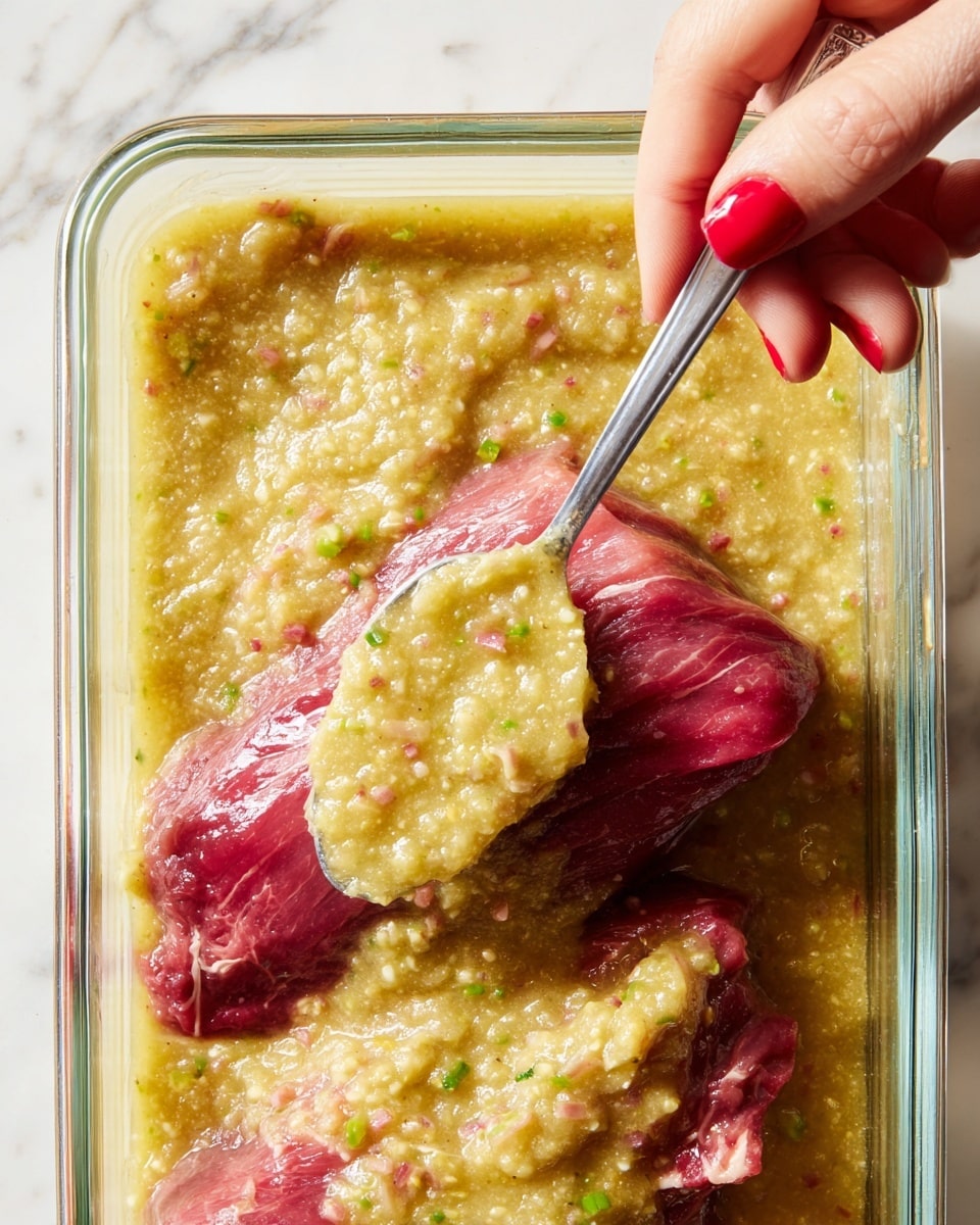 A clear glass container holds three pieces of raw red meat with visible marbling and texture, partially covered by a chunky, pale yellow sauce mixed with small green bits. A woman's hand with red nail polish holds a silver spoon spreading the sauce over the meat. The sauce has a thick, coarse texture with tiny pieces visible throughout, creating a contrast against the smooth meat underneath. The scene is set against a white marbled surface. photo taken with an iphone --ar 4:5 --v 7