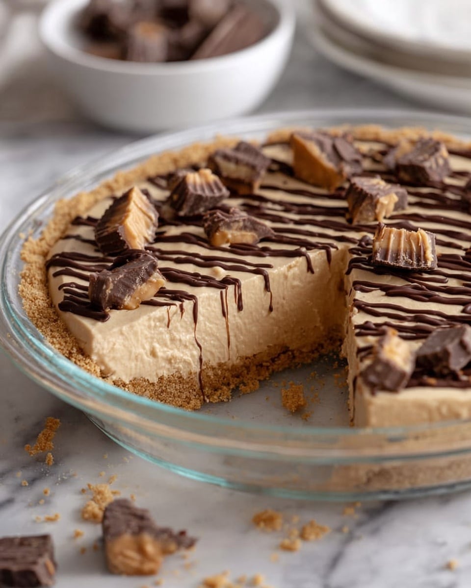 A pie with a crumbly light brown crust at the bottom and sides, filled with a thick, creamy tan layer in the middle. The top layer is decorated with dark brown chocolate drizzle in straight lines across the surface. On top, there are pieces of chopped chocolate peanut butter cups scattered evenly. The pie is placed in a clear glass pie dish on a white marbled surface, with some crumbs around the dish. In the background, a white bowl with more chocolate pieces is visible, softly blurred. photo taken with an iphone --ar 4:5 --v 7