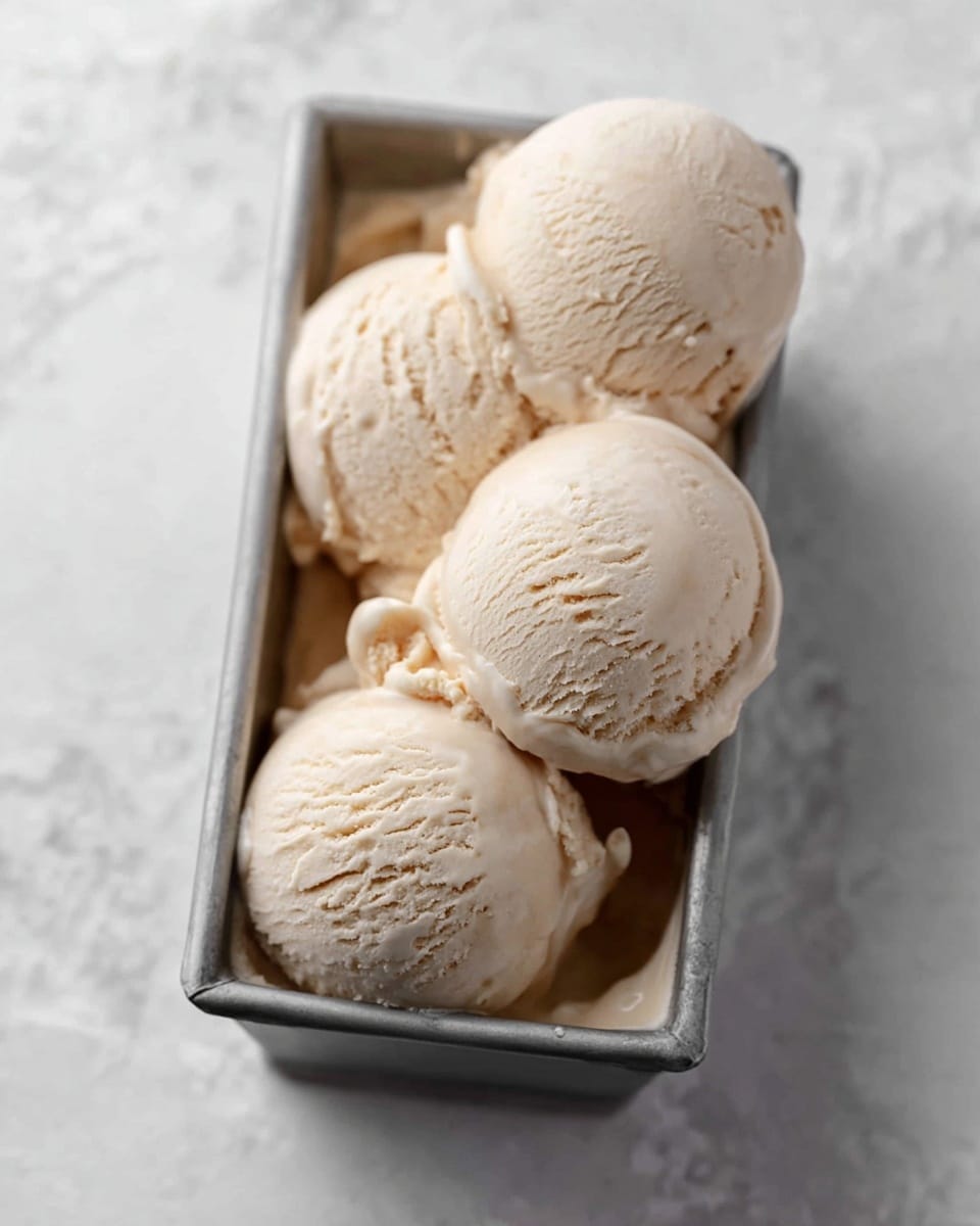 Four light beige ice cream scoops with a smooth and creamy texture are piled in a metal rectangular container, with some ice cream melted slightly at the base. The background is a white marbled texture that adds a clean and bright contrast. Photo taken with an iphone --ar 4:5 --v 7