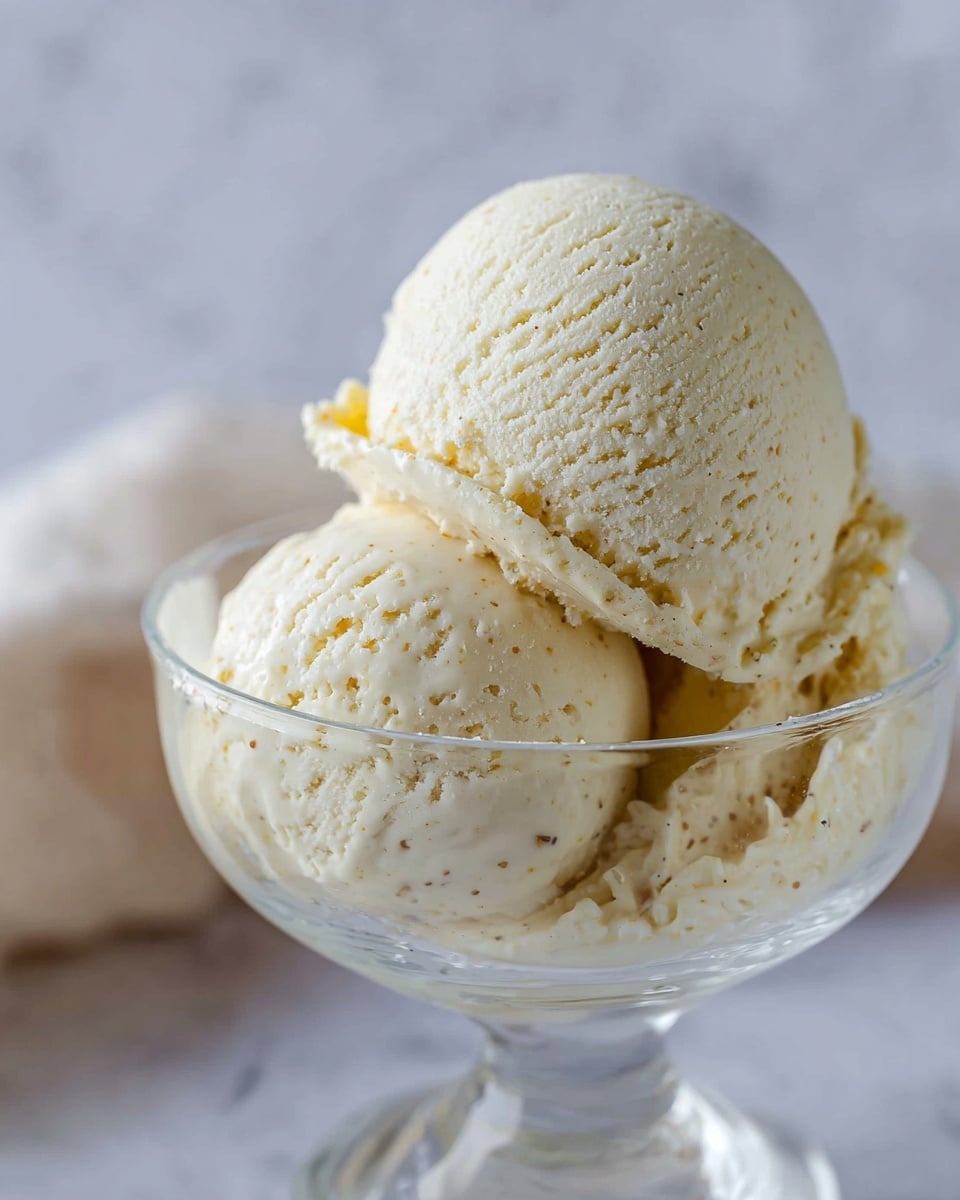 The image shows two scoops of creamy vanilla ice cream in a clear glass bowl. The top scoop is rounded and smooth with visible tiny specks of vanilla bean, while the scoop beneath is slightly rougher with a soft texture around the edges, creating a layered look. The clear glass bowl sits against a soft, blurred background with a white marbled texture underneath, giving a clean and simple feel. Photo taken with an iphone --ar 4:5 --v 7