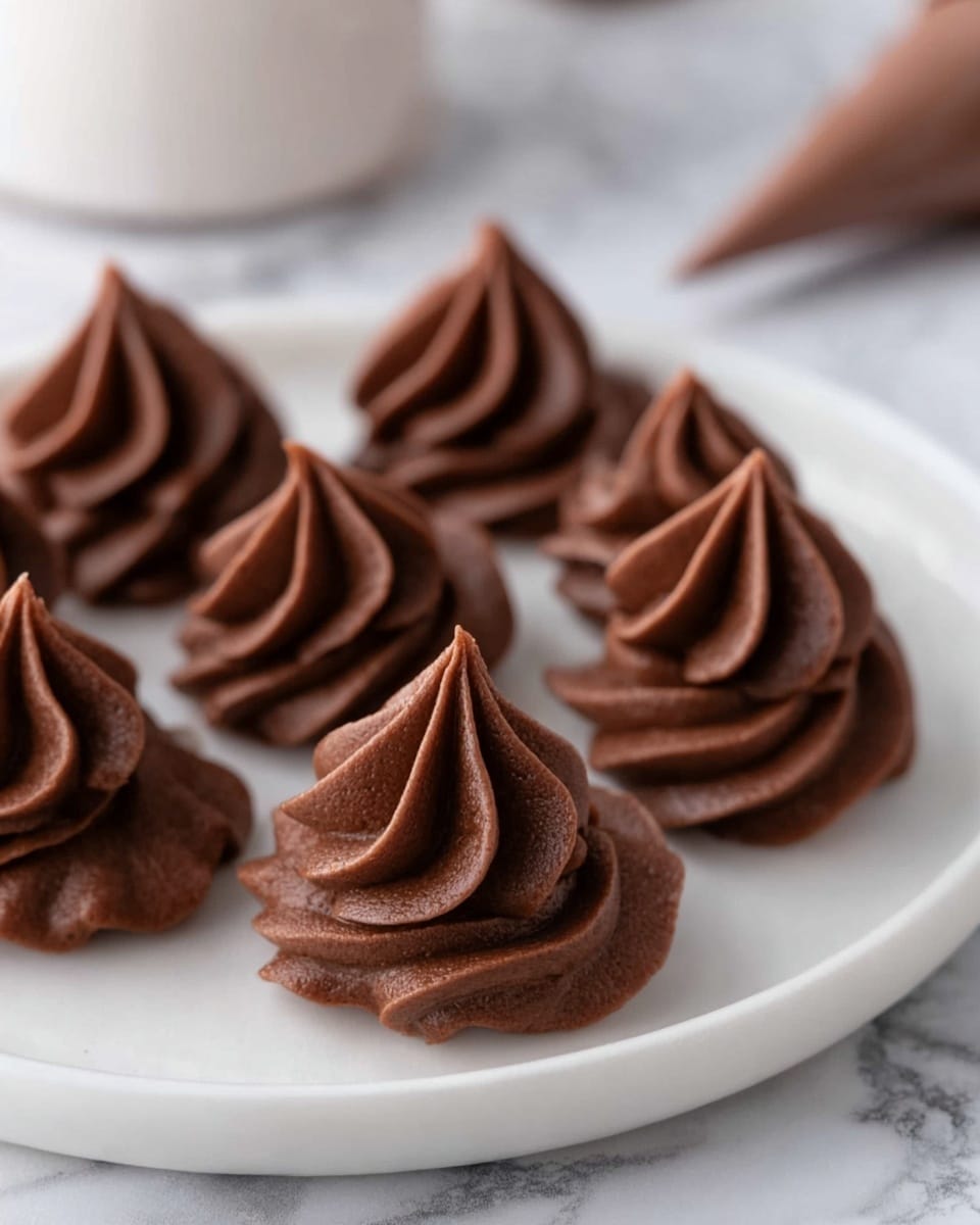 The image shows a white plate sitting on a white marbled surface, holding several dollops of chocolate frosting piped into small star shapes. The frosting has a smooth, creamy texture with a dark brown color, each dollop formed with multiple thick, curved layers that come to a point at the top. The shapes vary slightly but all share the same swirled, decorative style, arranged closely together on the plate. In the background, parts of a woman's hand and a piping bag are slightly blurred, suggesting the frosting is freshly piped. Photo taken with an iphone --ar 4:5 --v 7