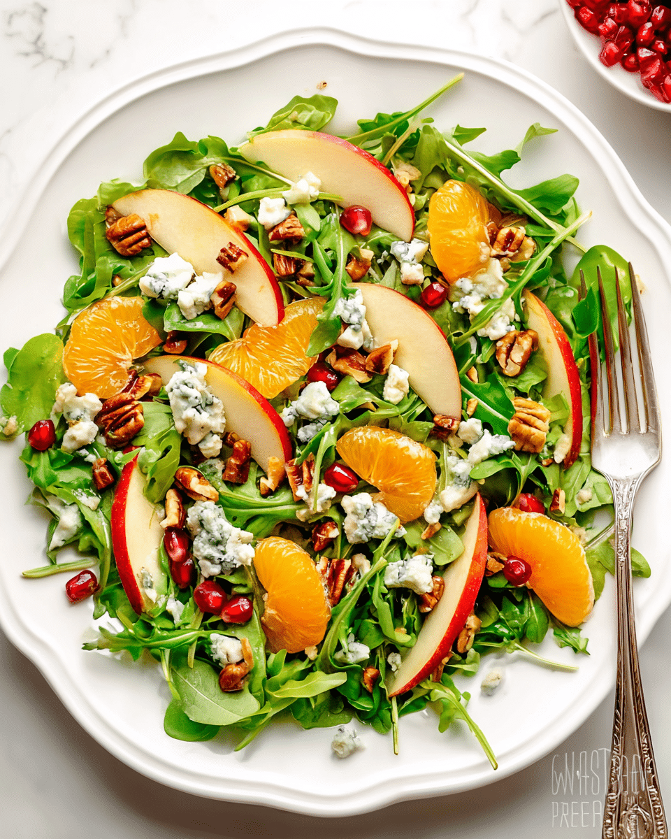 A fresh salad is arranged on a white plate with a scalloped edge, featuring a base layer of bright green arugula leaves with a slightly rough texture. On top are thin slices of red-skinned apple, tan with red edges, scattered evenly. Bright orange mandarin segments add a glossy, juicy look throughout. Small white chunks of blue cheese with blue veins are lightly sprinkled over the salad. Red pomegranate seeds add pops of red color, and small pieces of chopped brown pecans provide a crunchy texture. A silver fork rests on the right side of the plate against a white marbled surface. Photo taken with an iphone --ar 4:5 --v 7