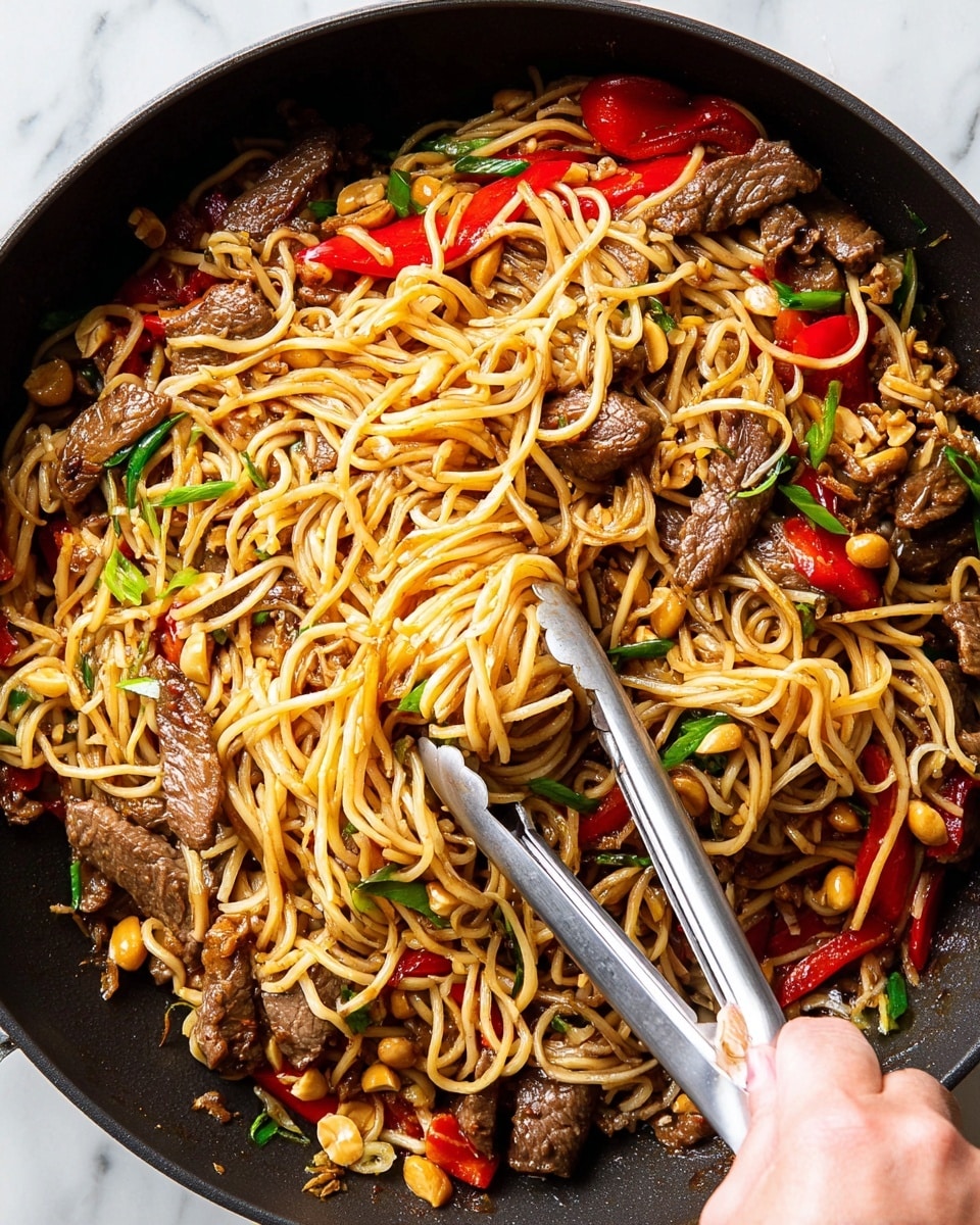 A close-up top view of a black pan filled with stir-fried noodles layered with thin brown beef strips, bright red bell pepper chunks, green onion slices, and golden peanuts scattered throughout. The noodles are light yellow and slightly shiny from the sauce, twisted and mixed with the other ingredients. A woman's hand is holding silver metal tongs that lift some noodles and beef from the middle, showing the mix's texture and color contrast. The background is a white marbled texture. photo taken with an iphone --ar 4:5 --v 7