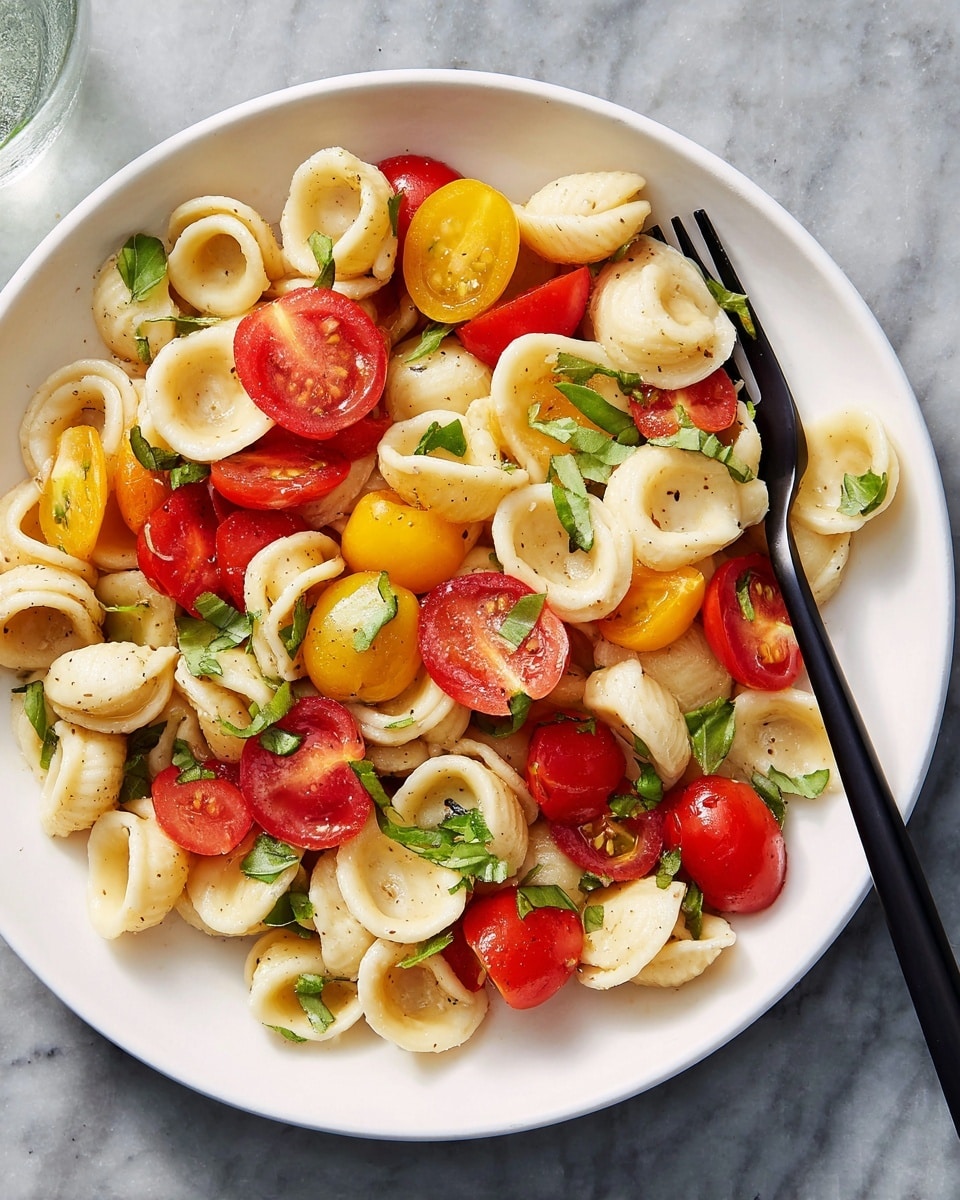 A white plate filled with a fresh orecchiette pasta salad layered with halved cherry tomatoes in bright red and yellow colors, scattered basil leaves adding green accents, and a light coating of oil that gives the pasta a slight shine. The pasta is cooked al dente, showing smooth round shapes with hollow centers. A black fork rests on the left side of the plate, spearing some pasta and a couple of tomato halves. The plate is placed on a white marbled surface. photo taken with an iphone --ar 4:5 --v 7